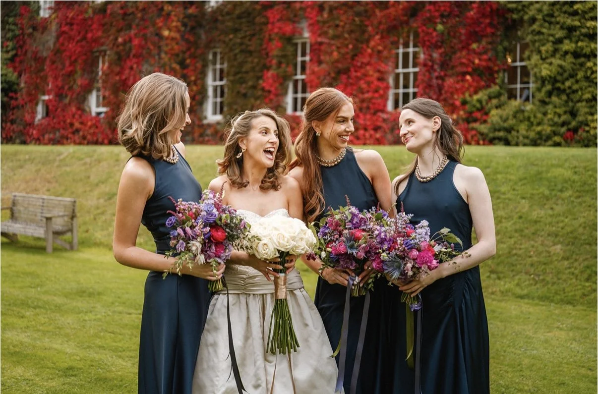 Four women at a wedding, three bridesmaids and a bride, standing outdoors on green grass in front of a building with red and green foliage. The bride is holding a bouquet of white roses. The bridesmaids are holding colourful bouquets