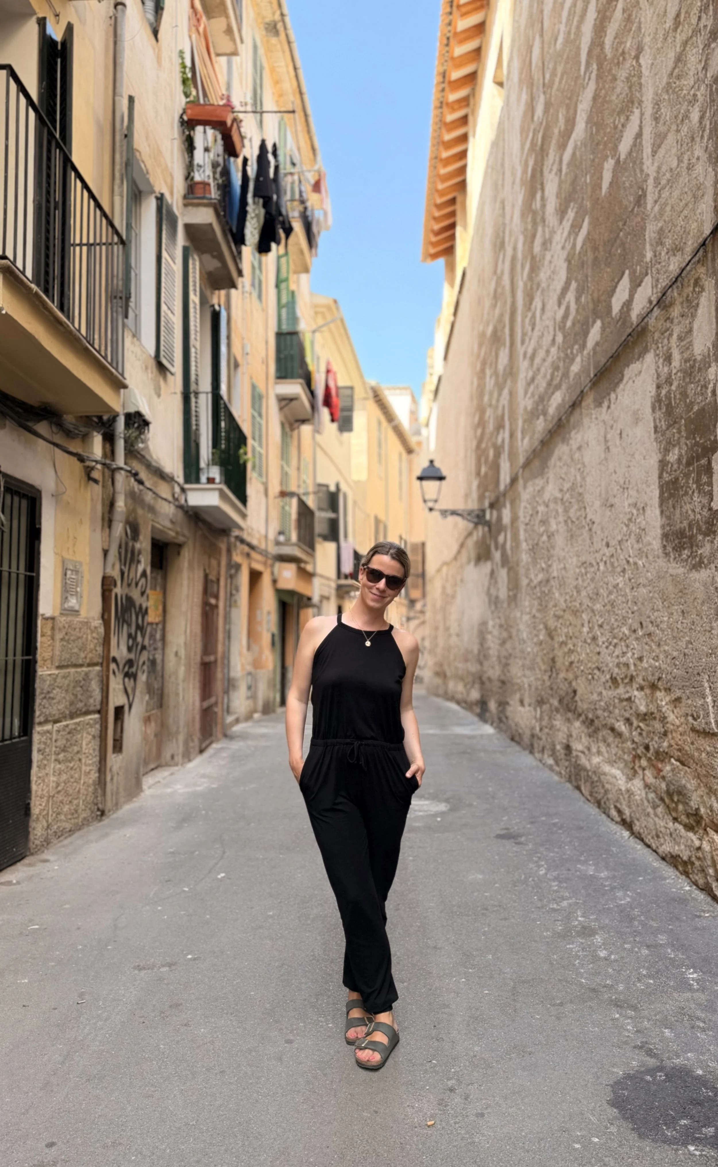 Eine junge Frau in einem schwarzen Sommeroutfit steht in einer engen, alten Gasse mit pastellfarbenen Gebäuden und Wäscheleinen. Die Frau trägt Sonnenbrille, Sandalen und lächelt.
