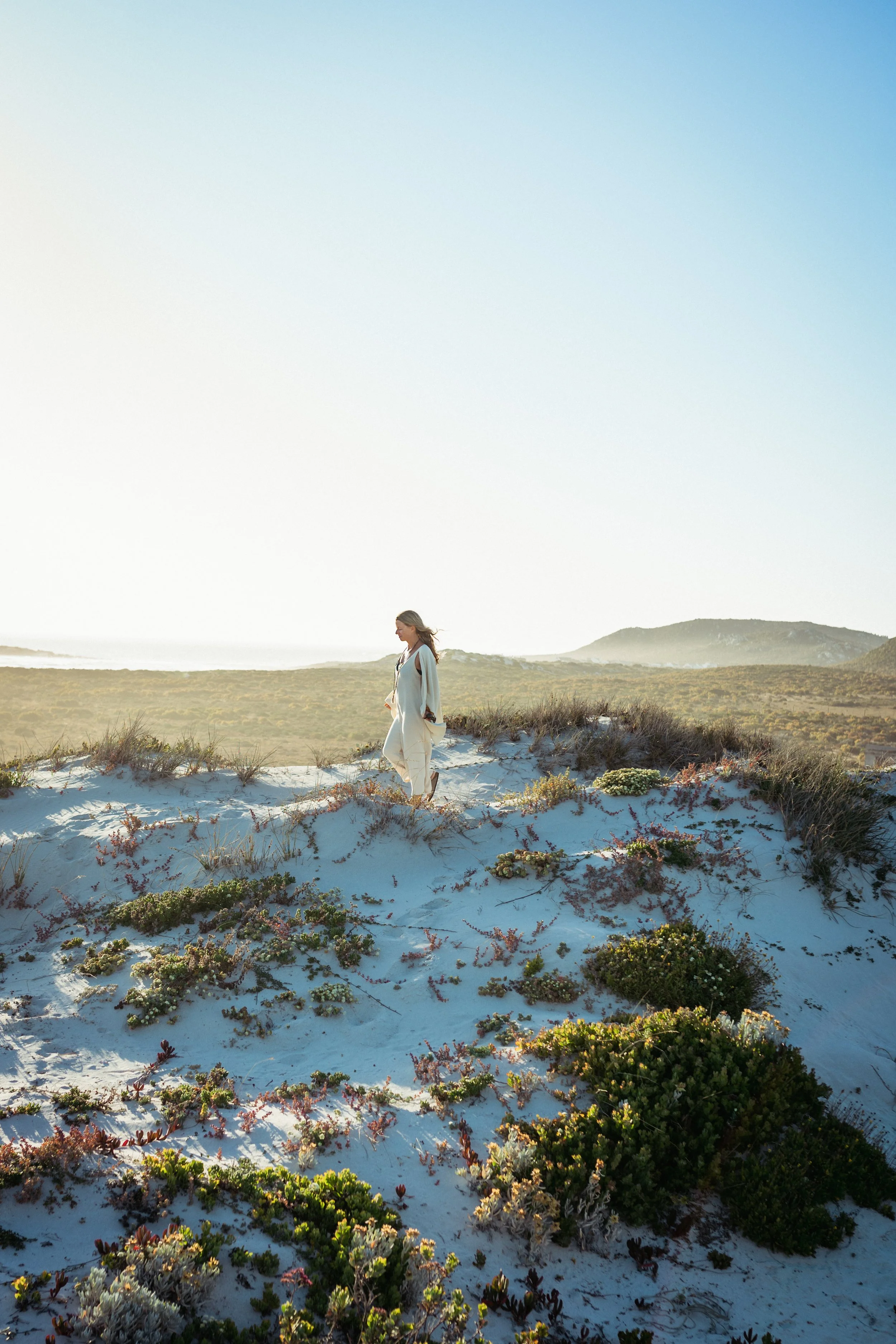 Eine Frau wandert auf Sanddünen mit Pflanzen, blauer Himmel und Berge im Hintergrund, Sonnenlicht, entspannte Atmosphäre.