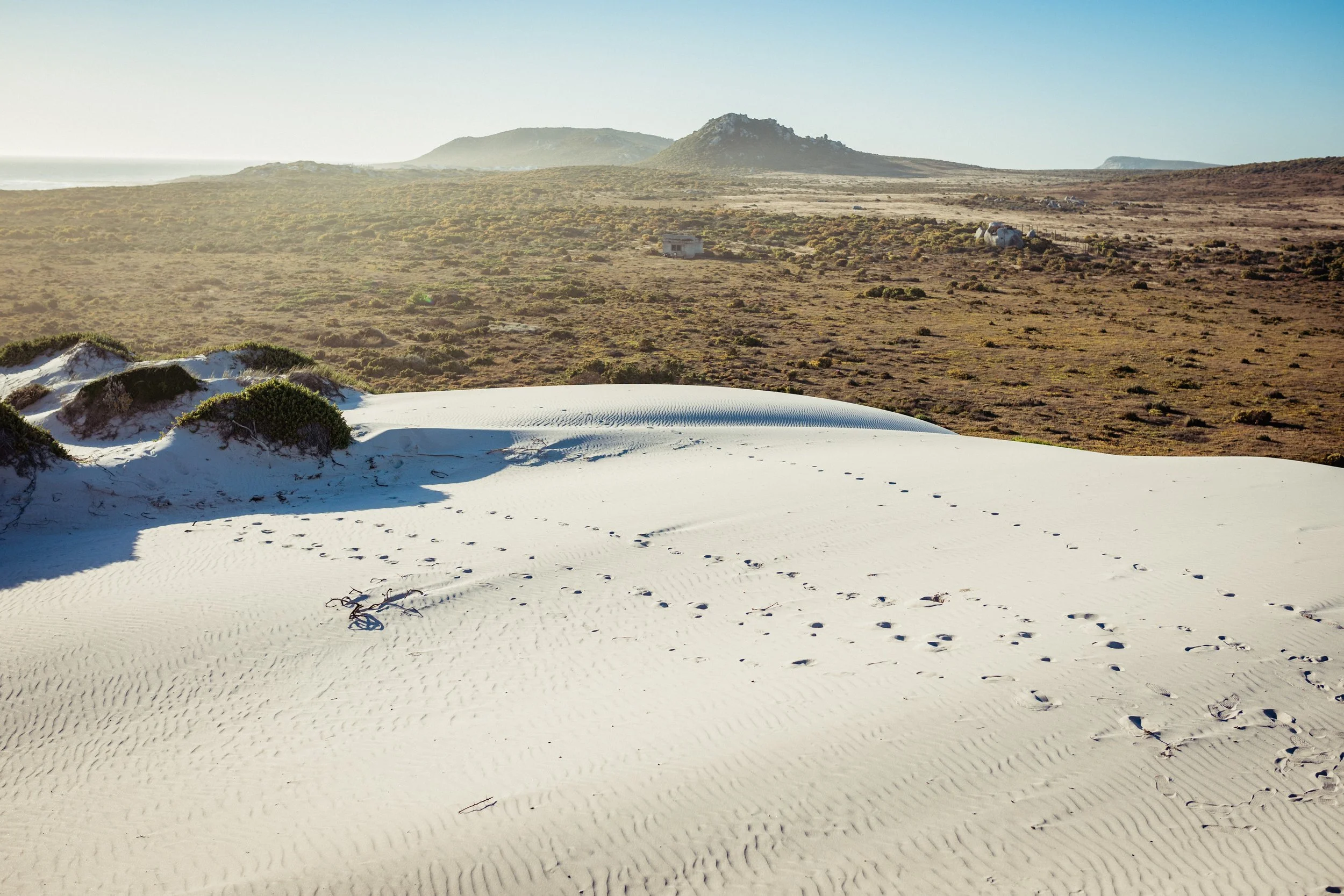 Sanddüne mit Schuhspuren und kleinen Sträuchern im Vordergrund, grasbewachsene Hügel und Berge im Hintergrund, blauer Himmel, sonnig.