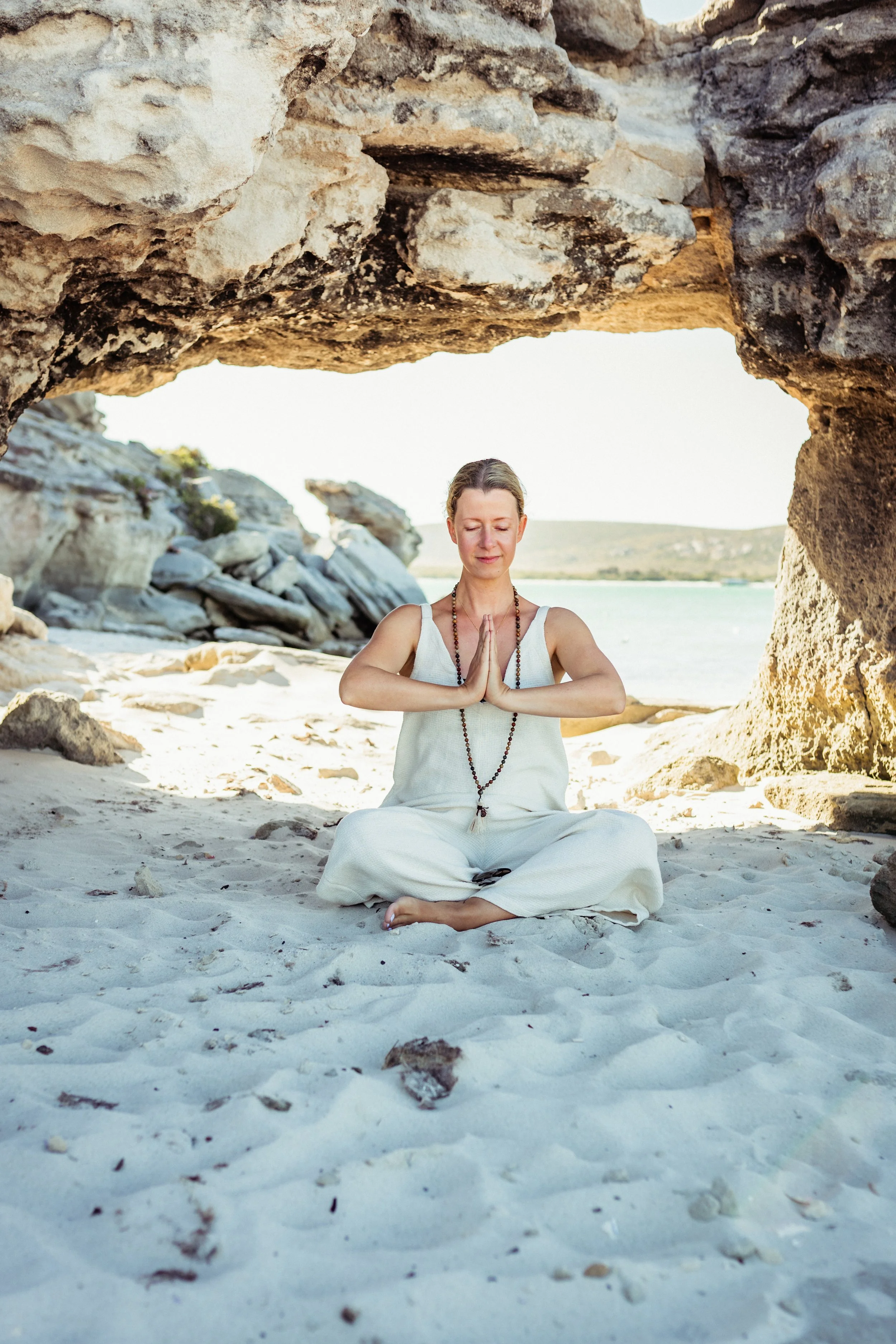 Eine Frau sitzt im Schneidersitz auf einem Sandstrand, meditiert mit geschlossenen Augen und betet die Hände vor ihrem Herzen. Sie trägt helle Kleidung, eine Halskette und eine Gebetskette. Im Hintergrund sind Felsen und das Wasser bei sonnigem Wetter.