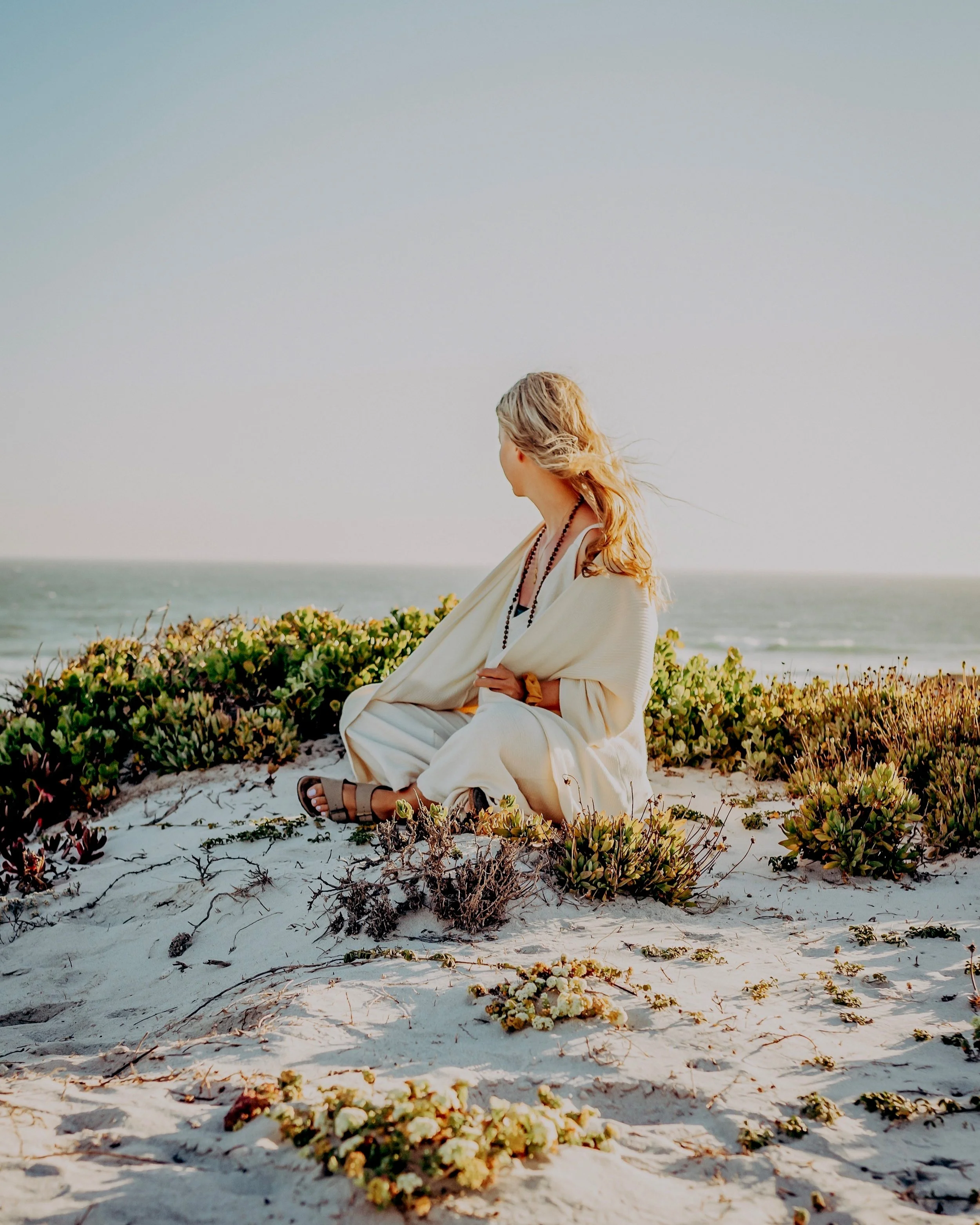 Frau sitzt im Sand am Strand, blickt aufs Meer, trägt ein weißes Kleidung, bei Sonnenuntergang oder -aufgang.