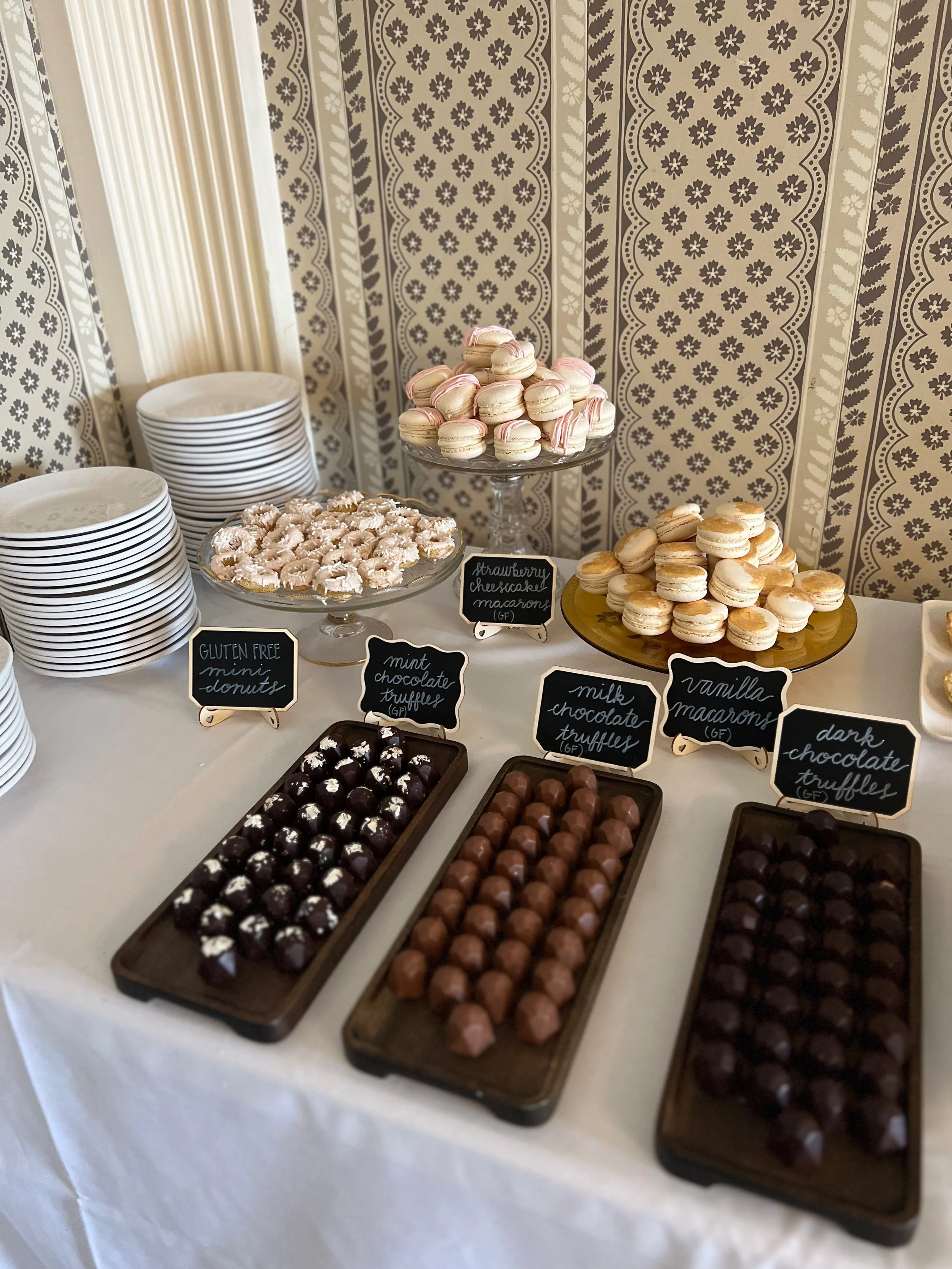 Assorted desserts on a table including mini donuts, macarons, and chocolate truffles with small black chalkboard signs labeling the treats.