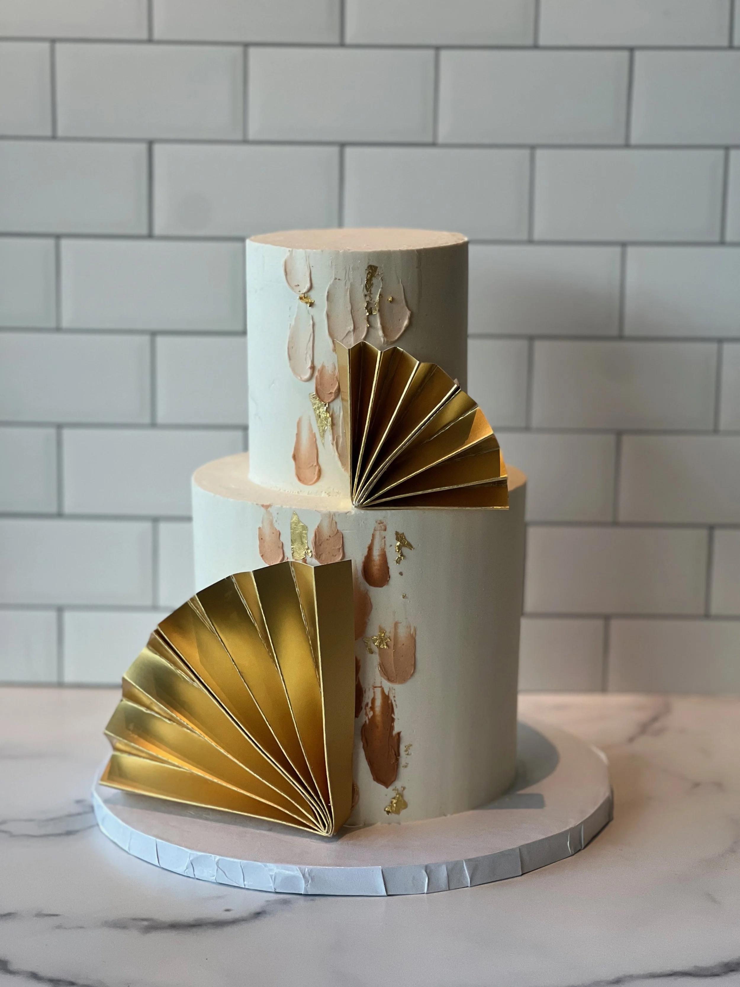 A two-tiered white cake with gold accents, decorated with pink and gold abstract drip patterns, topped with gold folded paper fans, placed on a white marble surface with a white tile wall in the background.