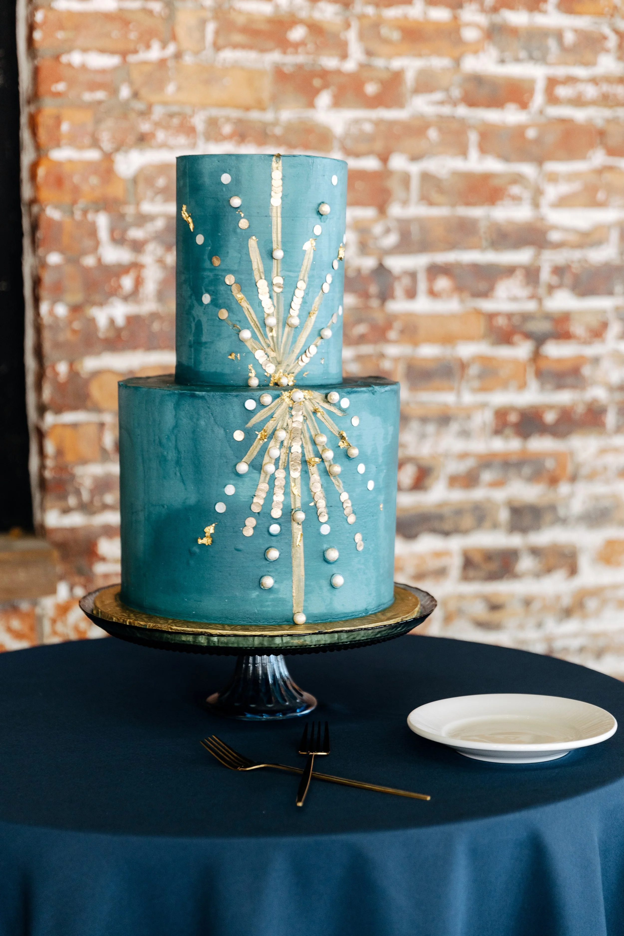 A two-tiered teal wedding cake decorated with white and gold accents, sitting on a black cake stand on a dark blue tablecloth with a white plate and black forks nearby, against a brick wall background.
