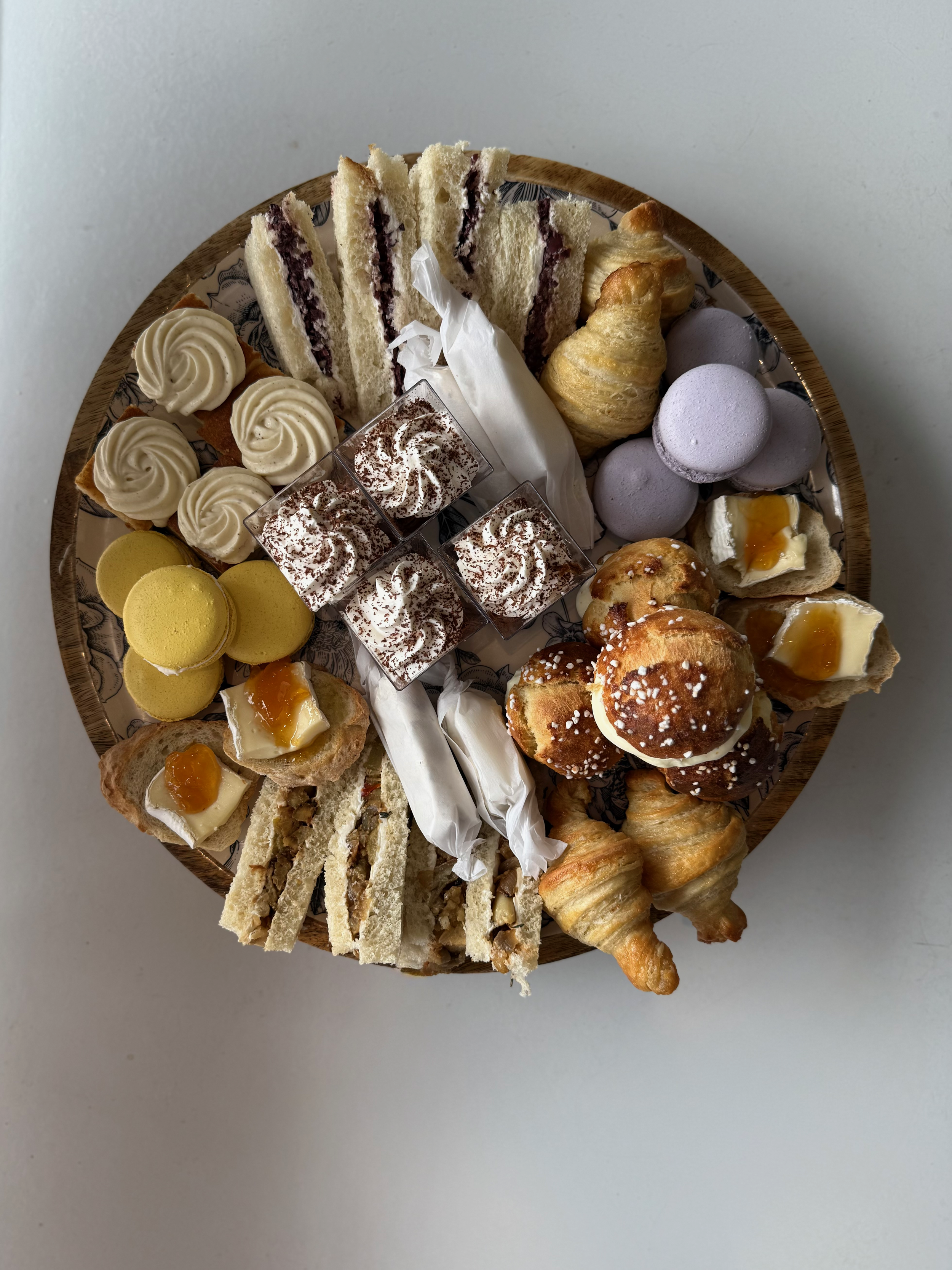Assorted desserts on a round wooden platter, including mini sandwiches with cream and jam, macarons, mini cakes, croissants, and profiteroles with frosting.