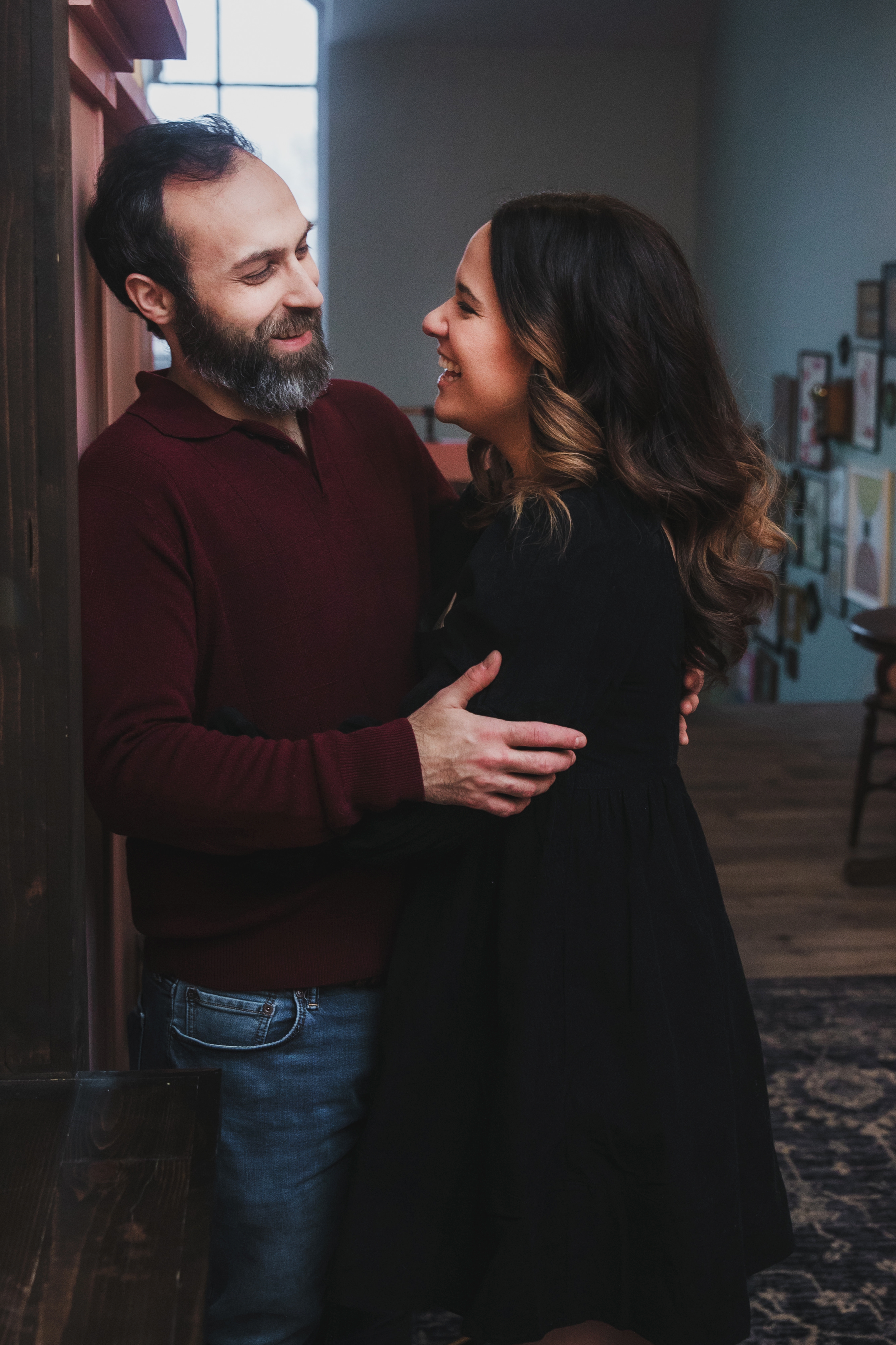 A man and woman share a joyful moment indoors, gazing at each other with smiles, standing close with their arms around each other.