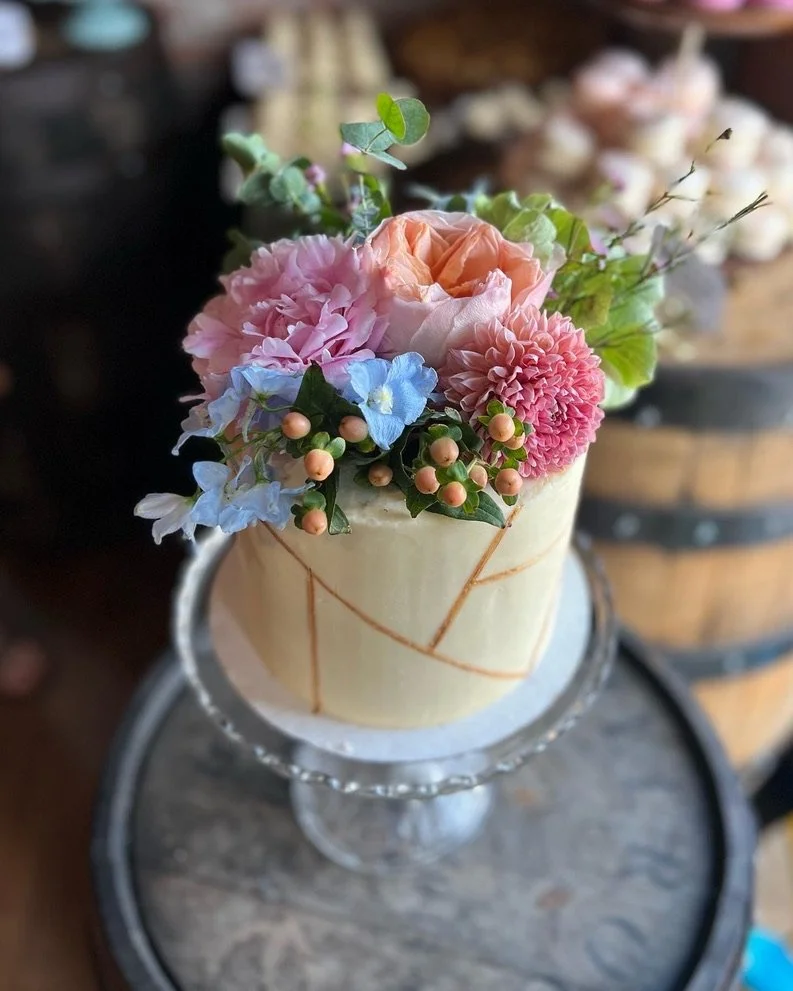 A floral cake on a glass cake stand with pink, peach, and blue flowers made of frosting or real flowers, topped with greenery.