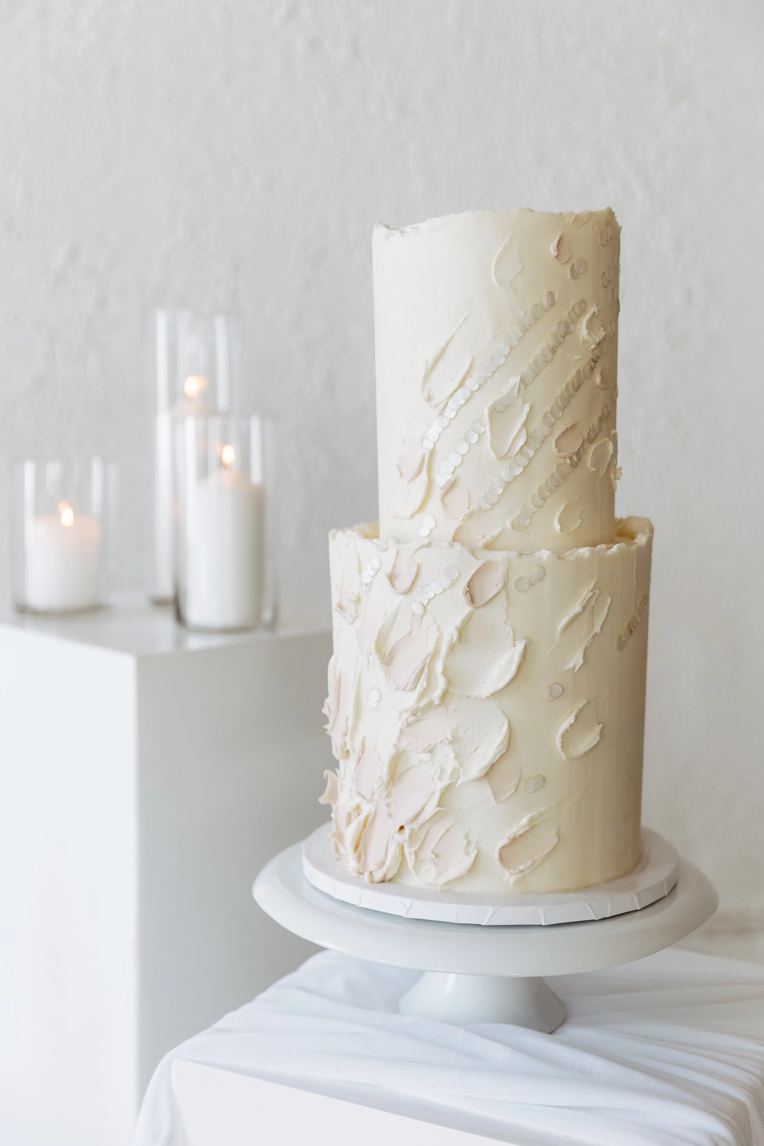 Elegant two-tier white wedding cake with textured icing and subtle pastel accents on a white cake stand, with lit candles in vases in the background