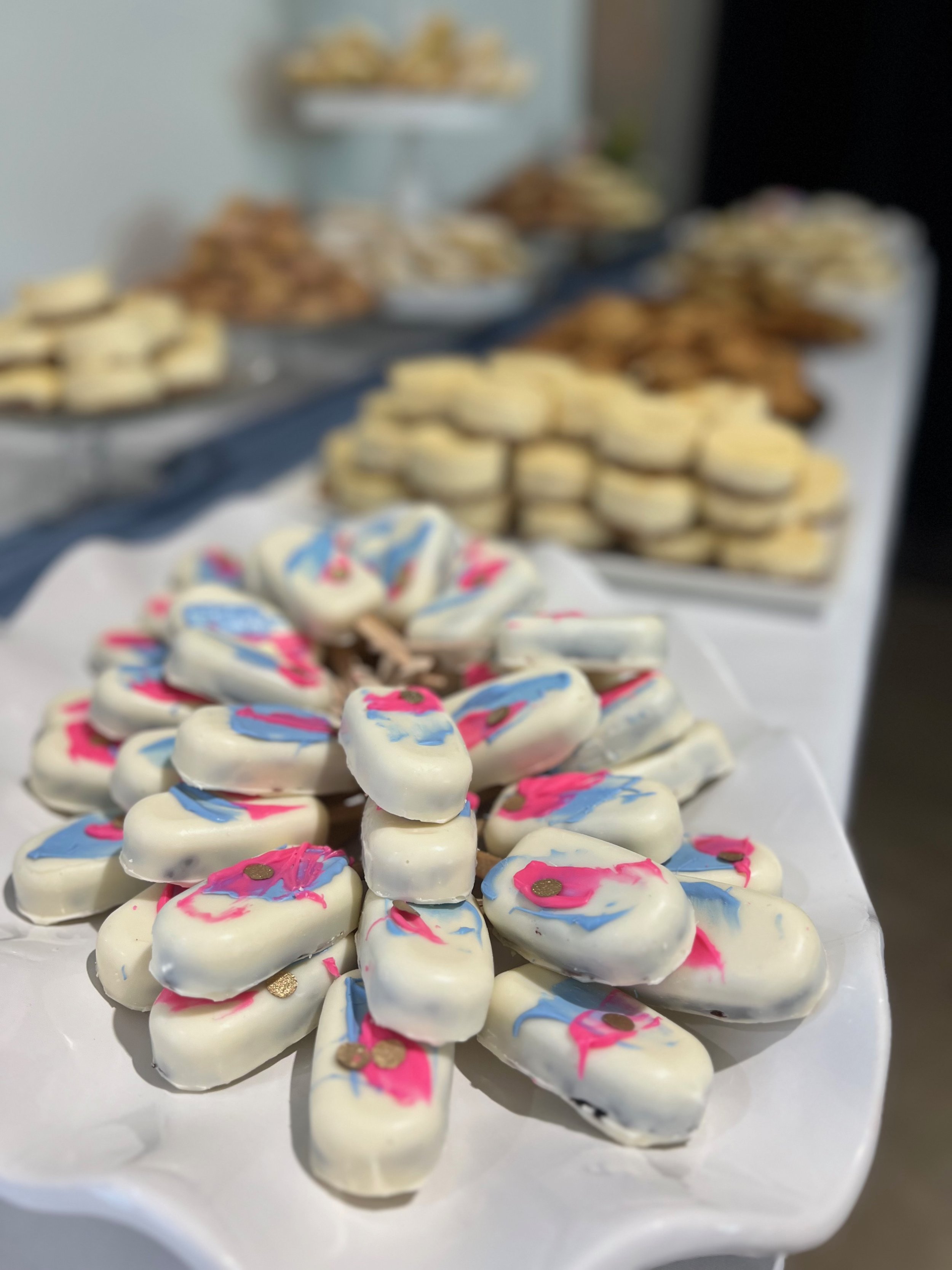 Tray of white candy coated almonds decorated with pink, blue, and gold accents, with more trays of cookies and desserts in the background.