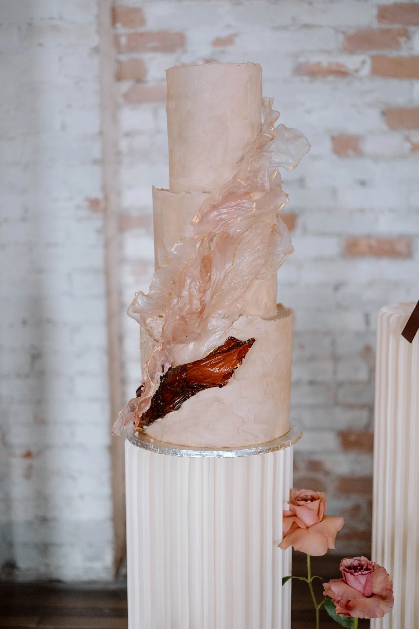 A tall, multi-tiered cake with a minimalist design on a white fluted pedestal, featuring soft peach-colored frosting, textured sugar decorations, and a dark brown decorative element, with pink roses at the base, against a brick wall background.