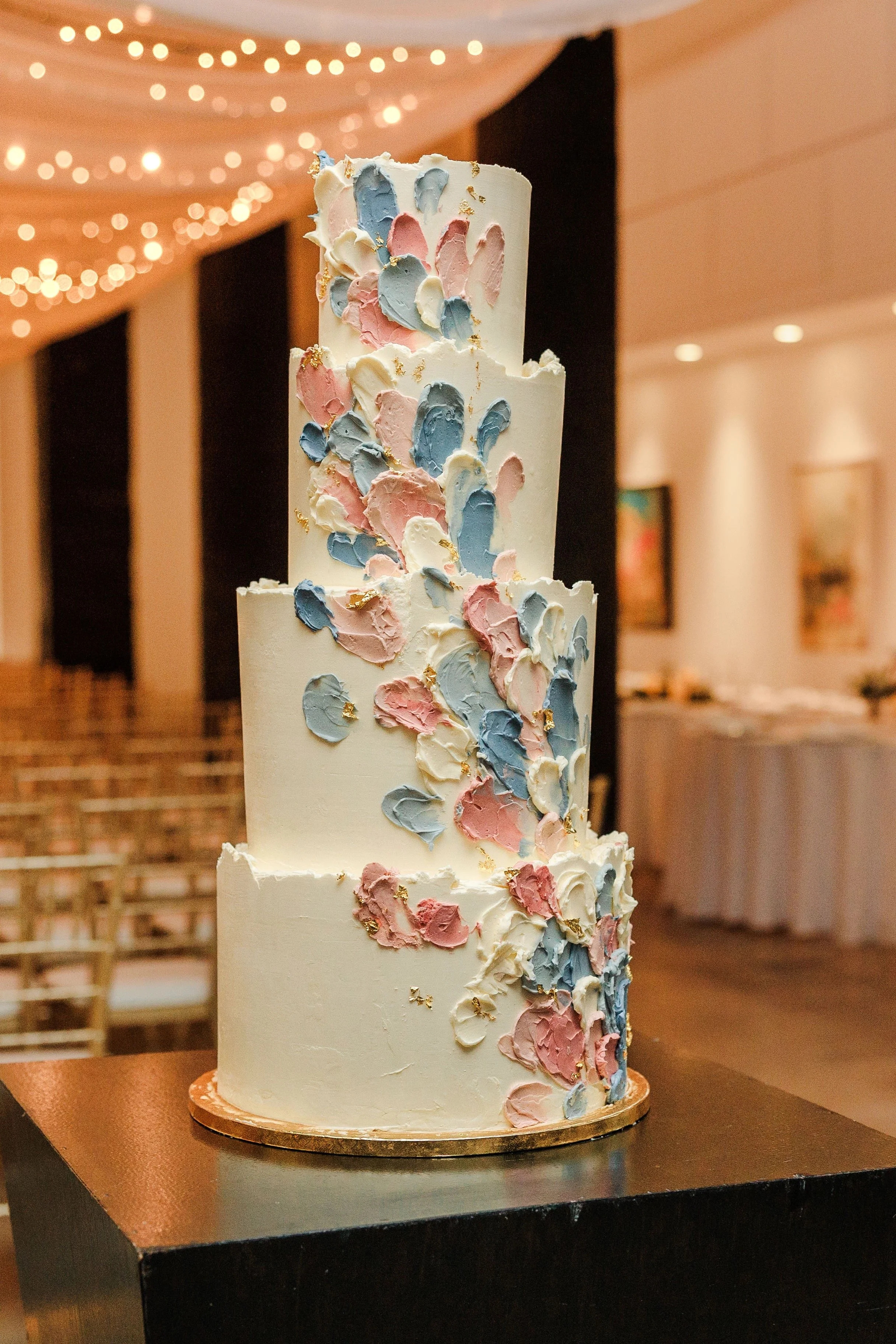 A tall, multi-tiered wedding cake decorated with pastel-colored buttercream swirls in pink, blue, and white, with gold accents, set in a decorated hall with string lights and artwork on the walls.