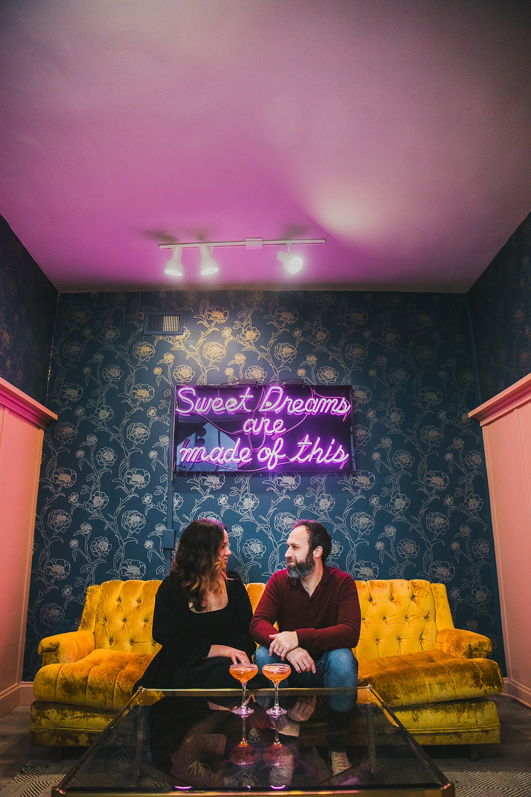A couple sitting on a yellow velvet sofa with two cocktails on a glass coffee table in front of them. Behind them, a dark floral wallpaper and a neon sign that reads 'Sweet Dreams are made of this' in pink and purple.