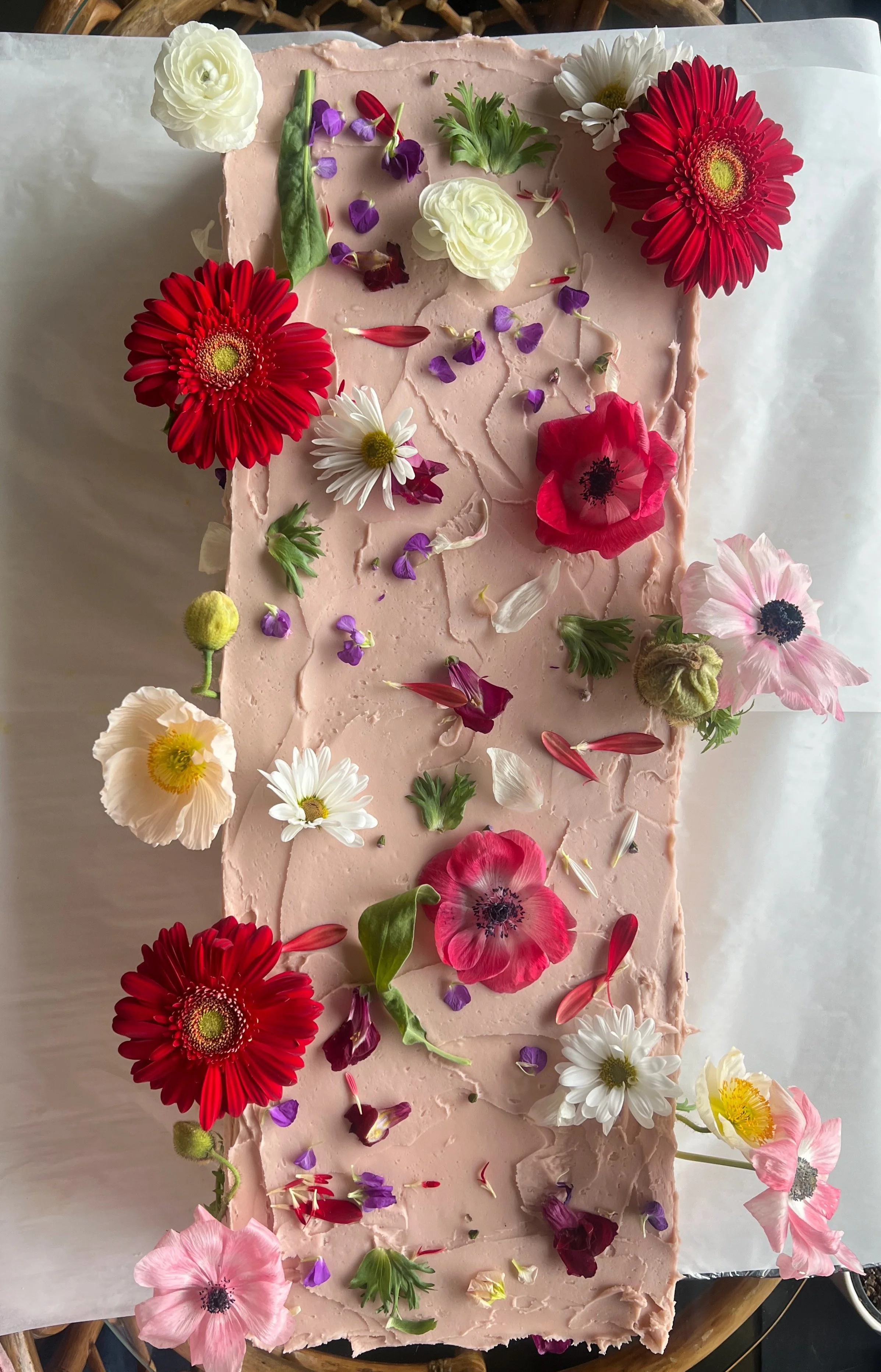 A rectangular pink cake decorated with fresh flowers, including red, white, yellow, pink, purple, and green flowers and petals, arranged on top and around the edges.