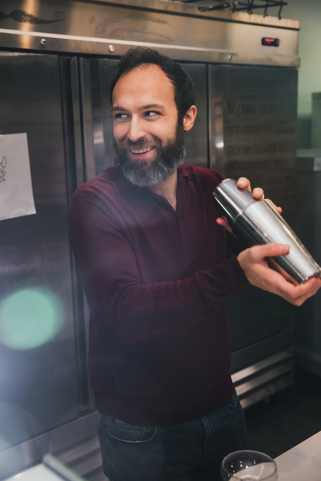 A man with a beard and dark hair, wearing a maroon long-sleeve shirt, smiling and holding a silver cocktail shaker in a kitchen.