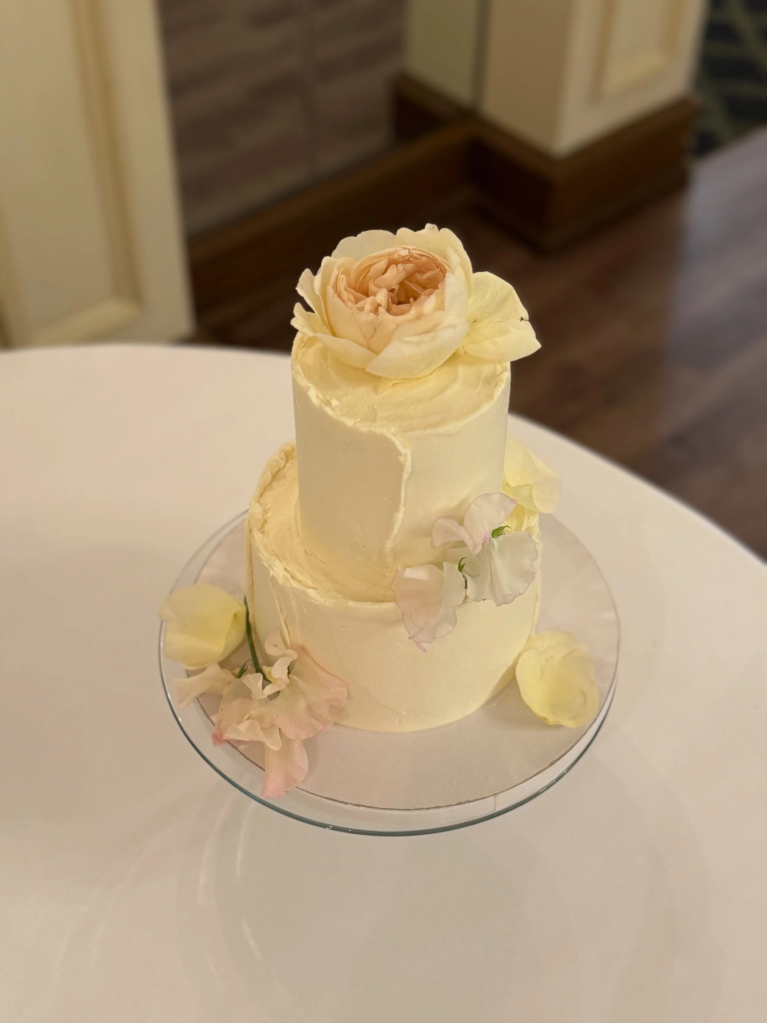 Two-tier white wedding cake decorated with fresh pink and white flowers on a glass cake stand.