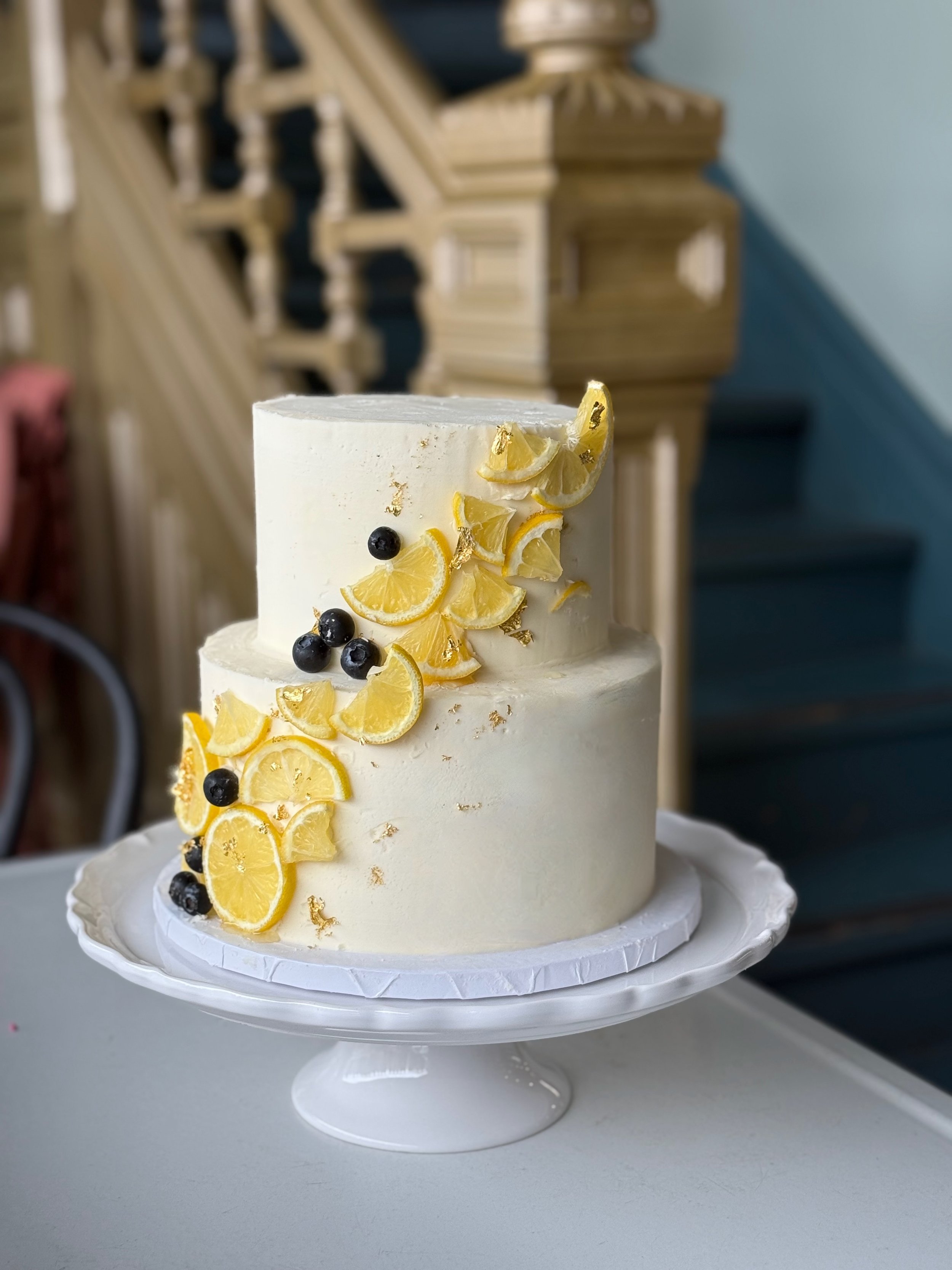 A two-tiered white cake decorated with lemon slices, black berries, and gold leaf on a white cake stand.