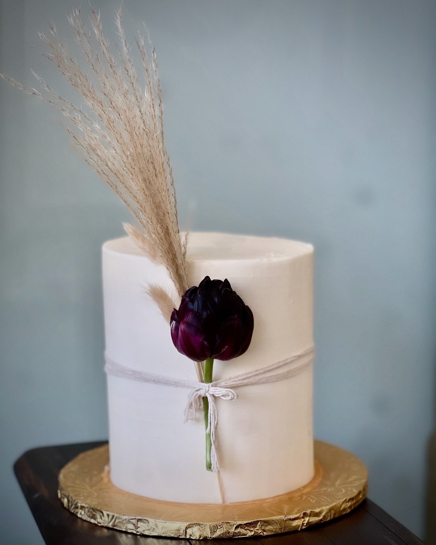 A white cylindrical cake with a black flower, beige dried grass, and tied with a twine bow, placed on a wooden cake board.