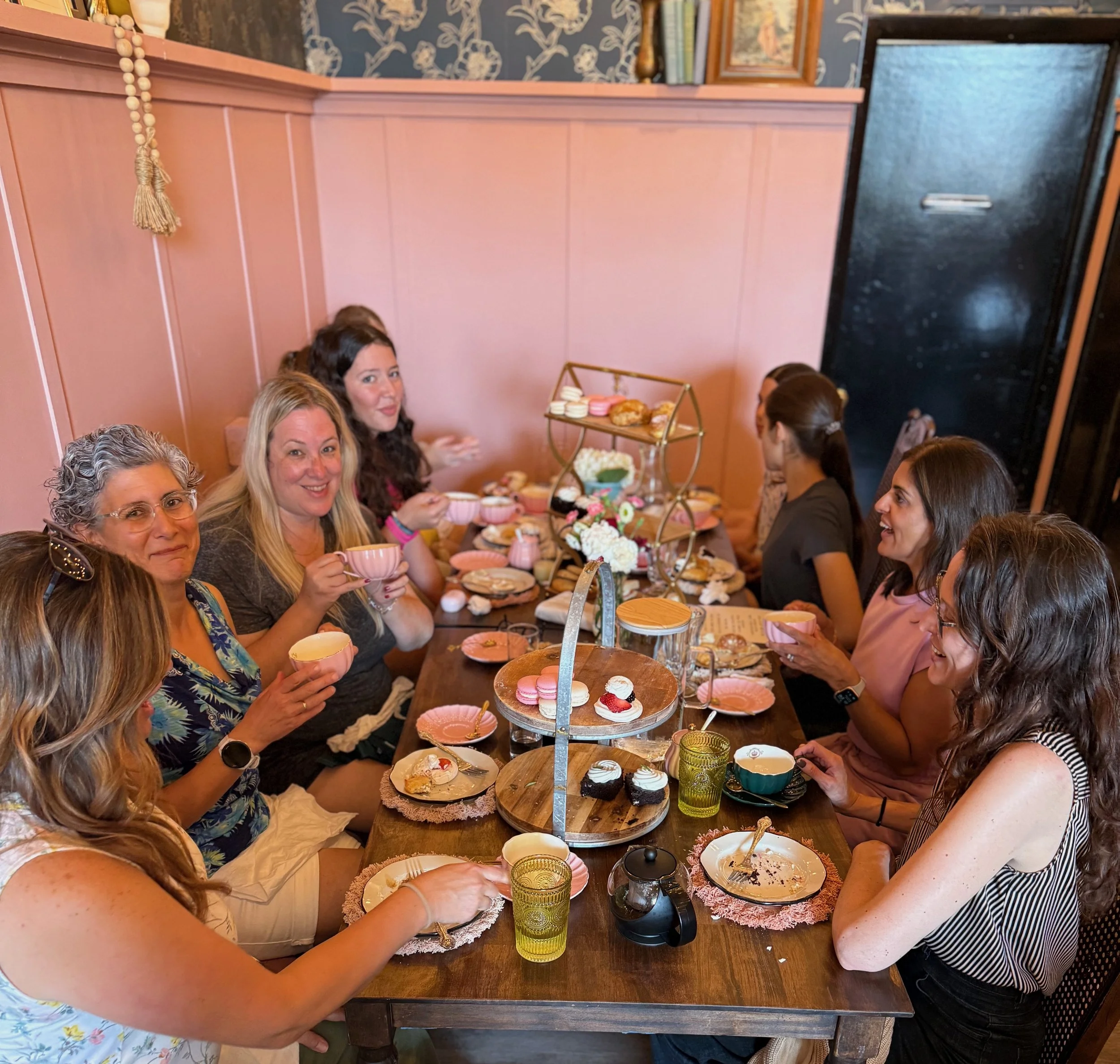A group of seven women enjoying a tea party in a cozy room, with a table filled with cupcakes, cookies, and tea cups. They are smiling and engaging in conversation.