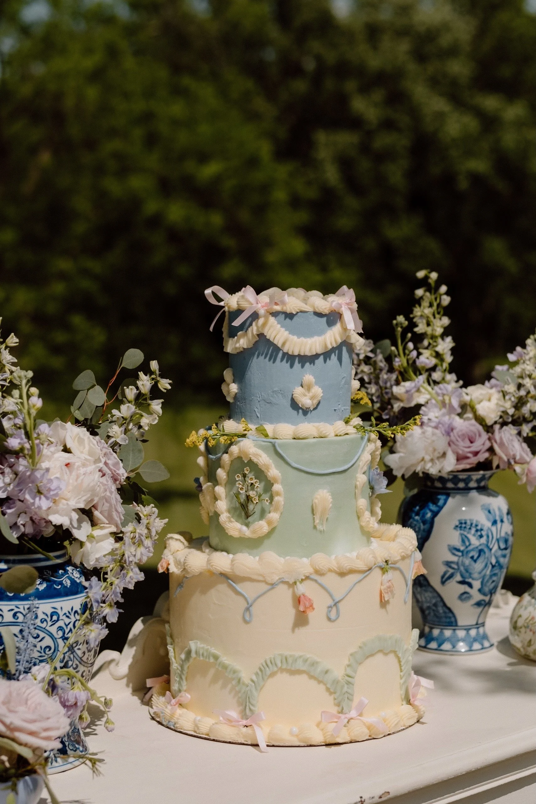 Three-tiered pastel-colored wedding cake decorated with white piped icing and small pink bows, surrounded by floral arrangements in blue and white vases.