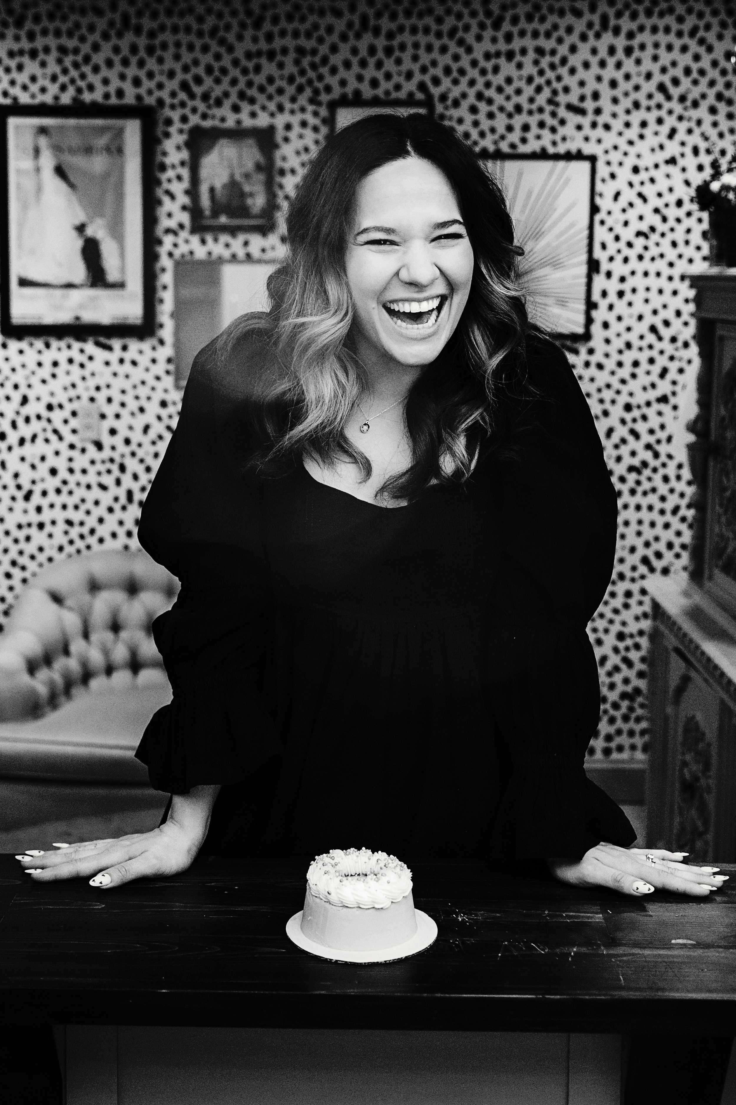 A woman with wavy hair laughing and leaning on a table with a small birthday cake, in a room with patterned wallpaper and framed pictures.