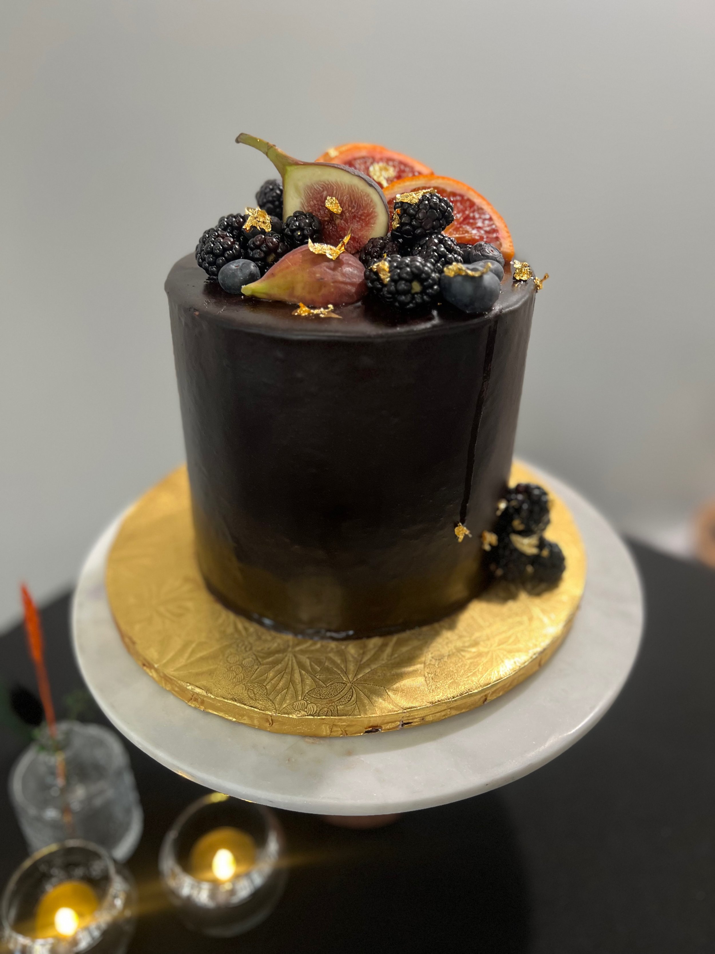 A tall, cylindrical chocolate cake decorated with fresh berries, figs, and edible gold leaves, displayed on a white cake stand with a gold-colored base.