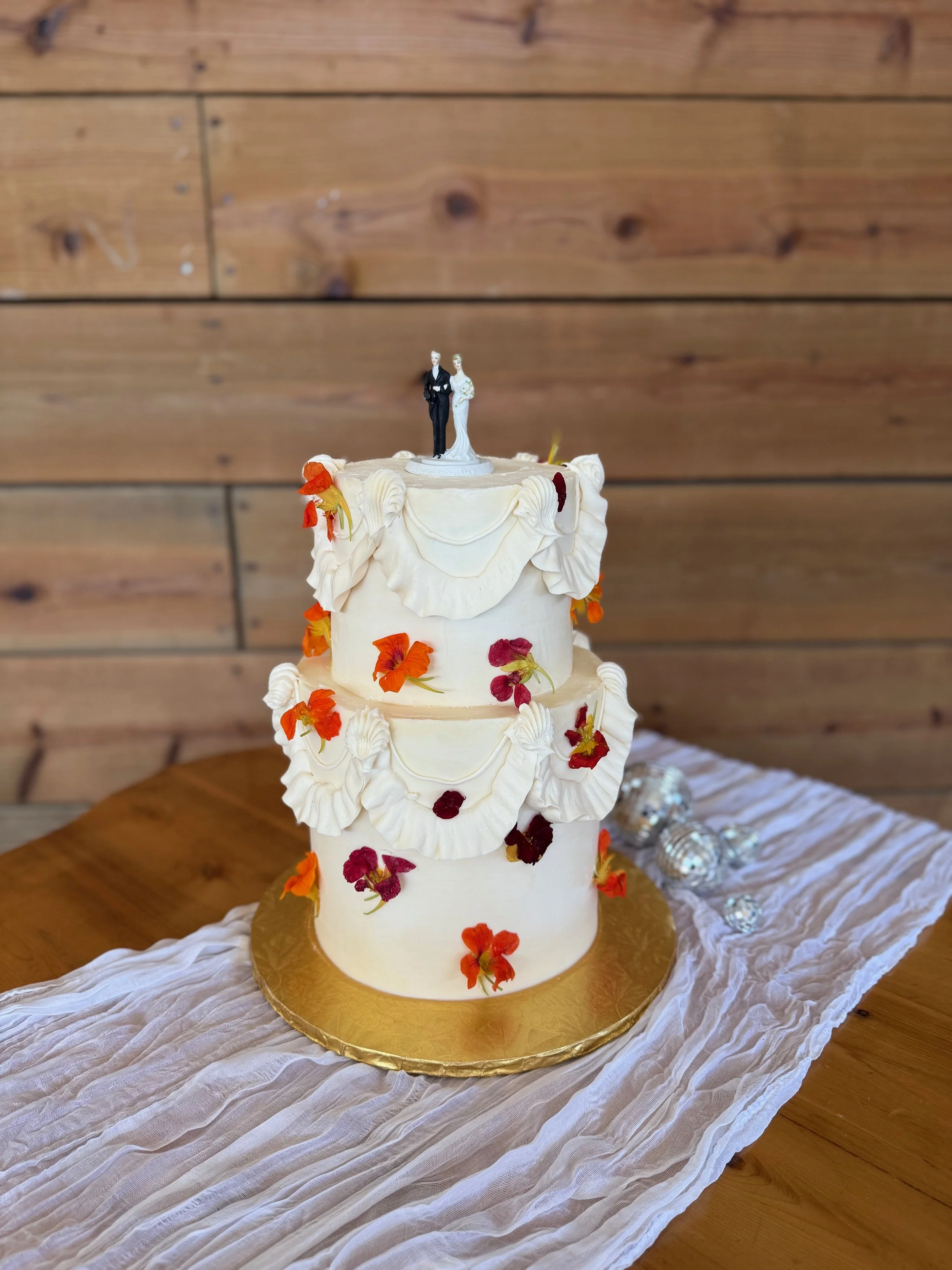 Three-tier wedding cake decorated with white icing, colorful edible flowers, and a bride and groom figurine on top, placed on a golden base on a table with a white cloth against a wooden wall background.