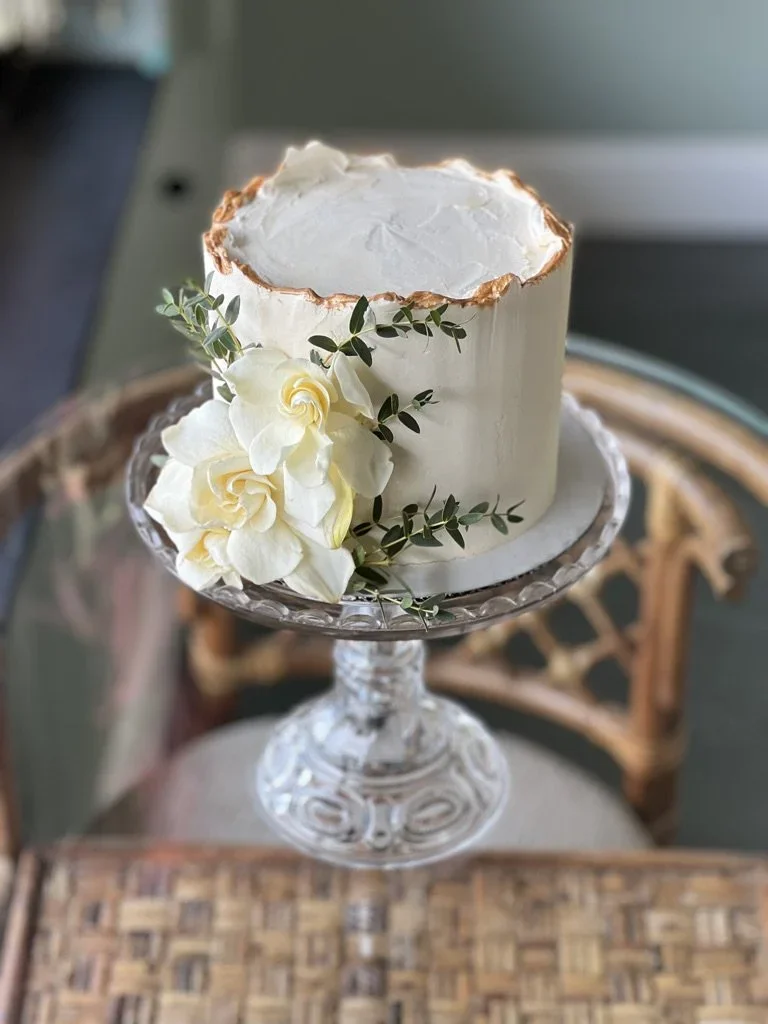 A tall white frosted cake with a gold-rimmed top, decorated with white flowers and green leaves, displayed on a glass cake stand.