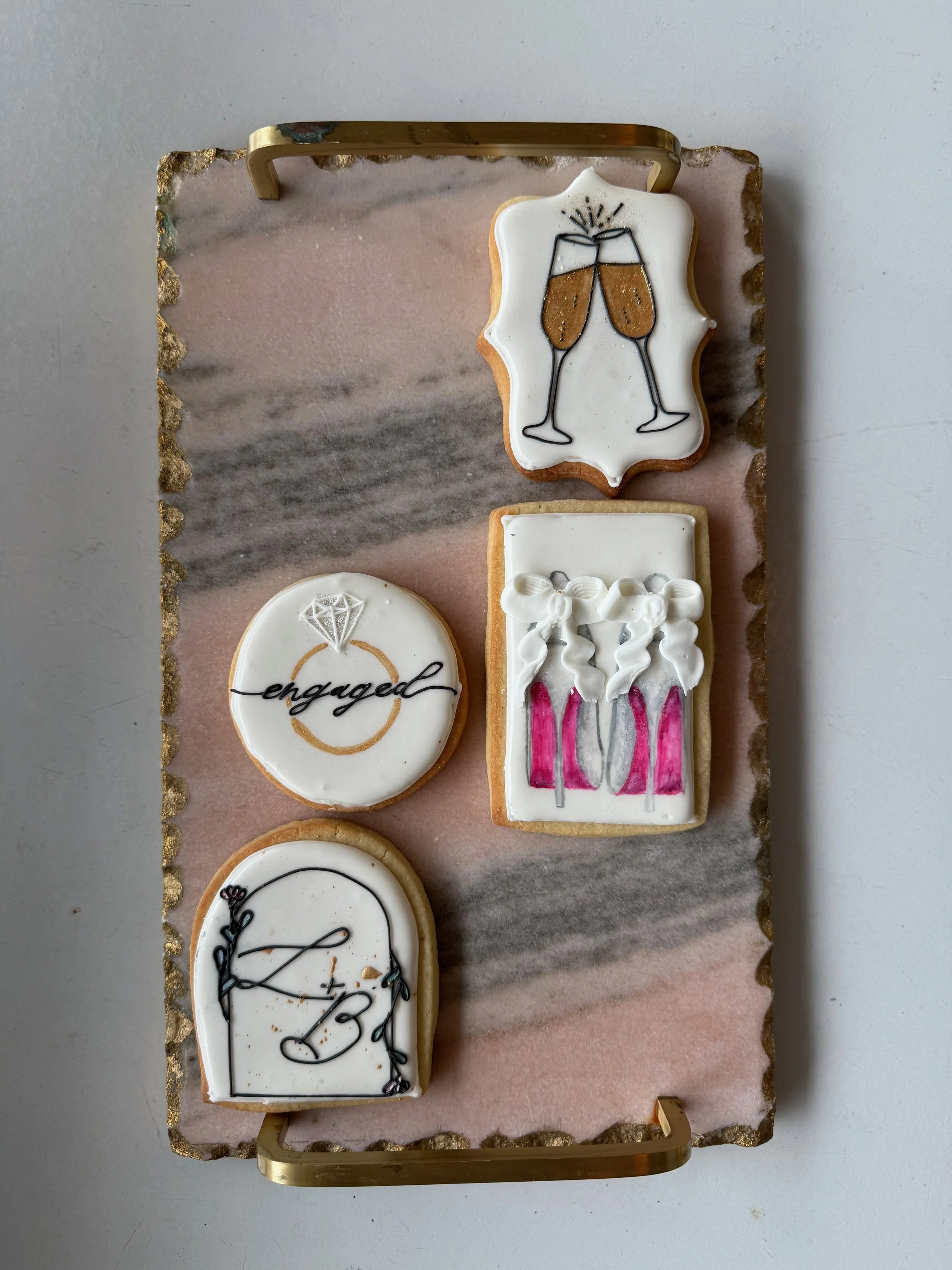 Set of five decorated cookies celebrating an engagement, featuring champagne glasses, a diamond ring with the word 'engaged,' balloons with 'I Do' along with hearts and pink ribbons, and a blank plaque with decorative elements, arranged on a pink and gray marble tray with gold trim and handles.