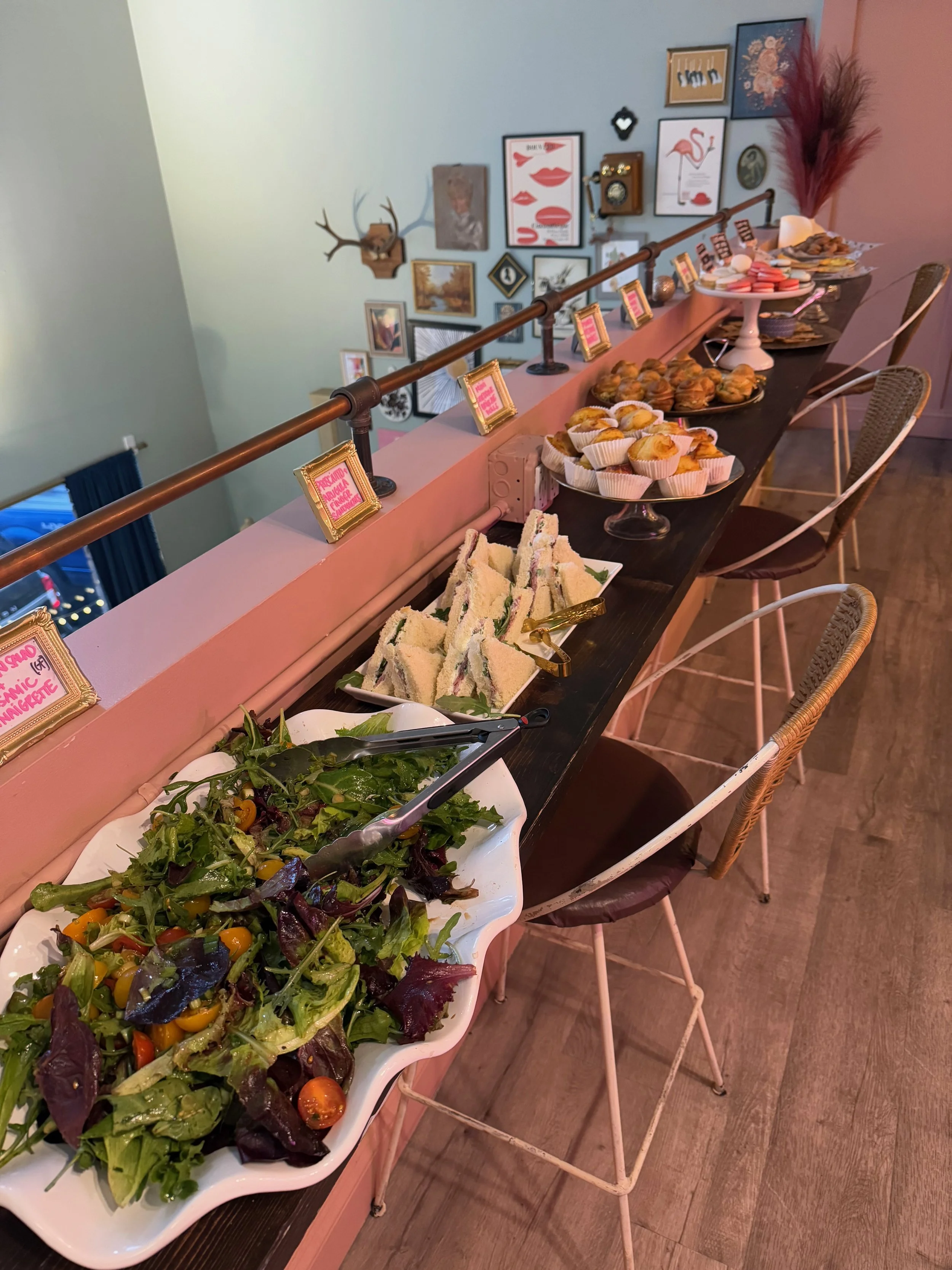 A buffet table with assorted salads, sandwiches, muffins, and desserts, set against a wall decorated with various framed pictures and artwork.