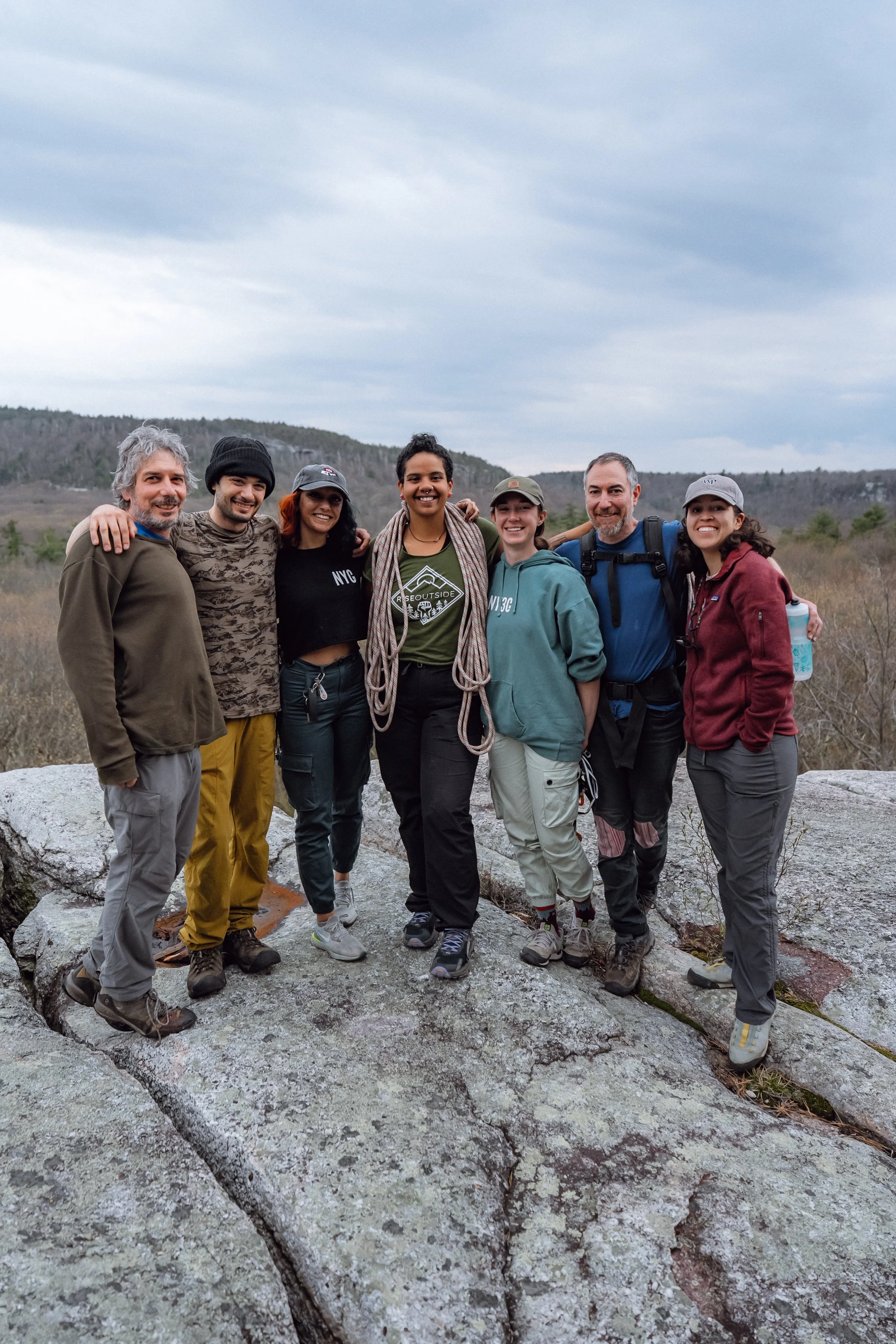 Seven climbers stand arm-in-arm on a rocky overlook during an outdoor climbing trip, smiling with a forested landscape behind them.