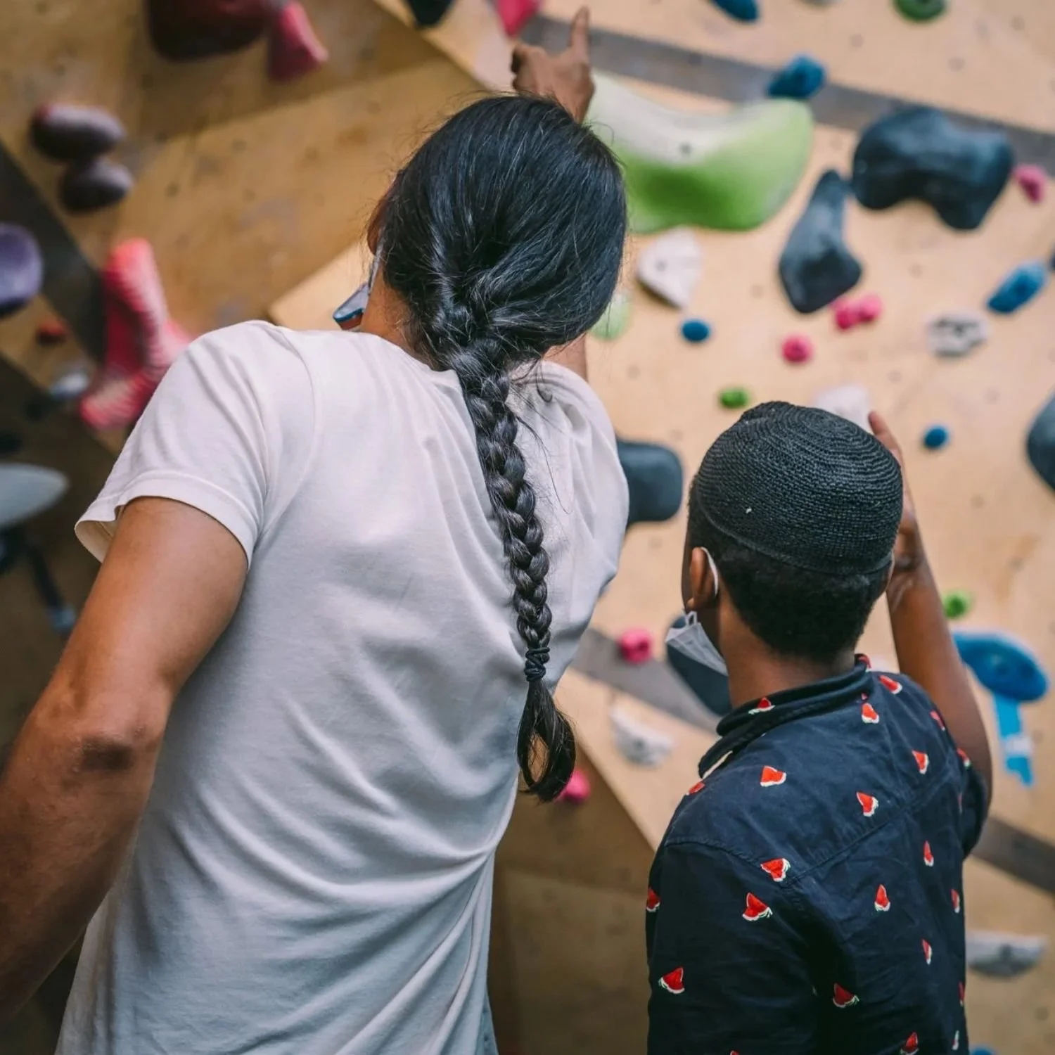 A mentor with a braided ponytail points to an indoor climbing route for a child.