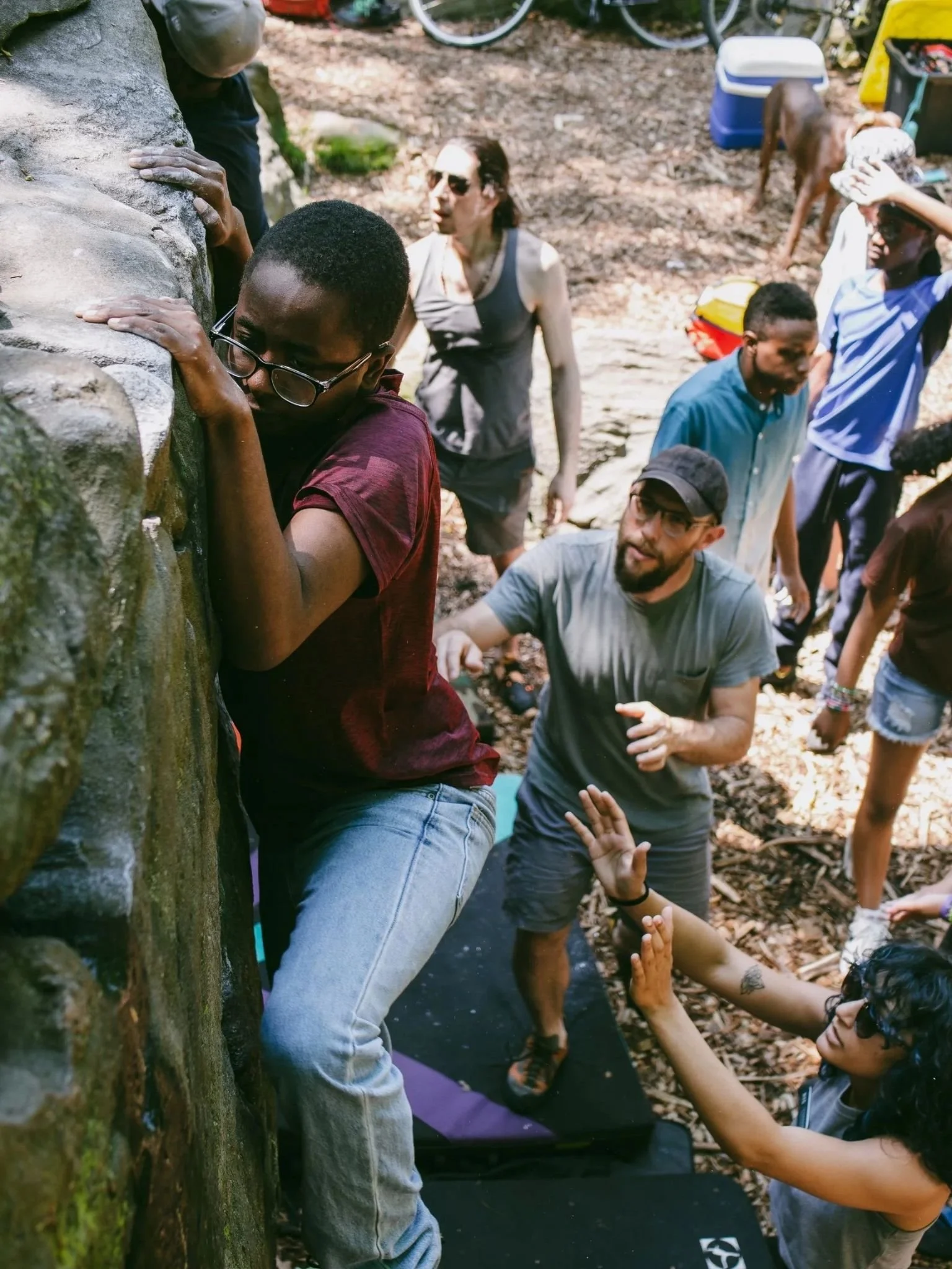 A student climbs a rock face while peers and mentors spot from below.