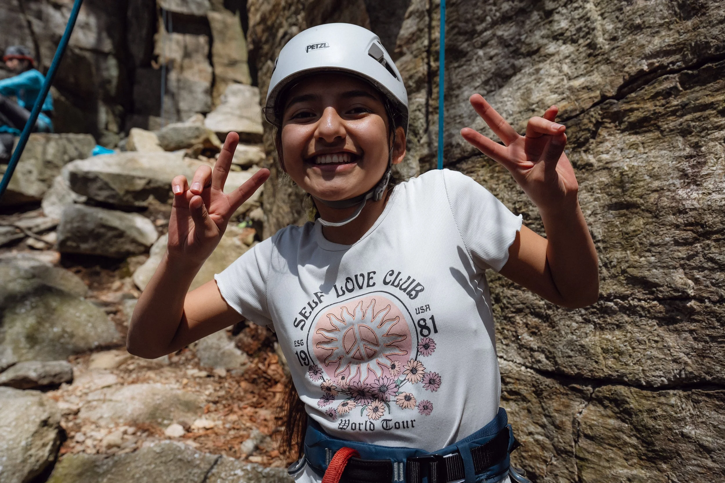 A smiling young climber wearing a white helmet and harness flashes peace signs while standing at the base of a rocky cliff during an outdoor climbing activity.