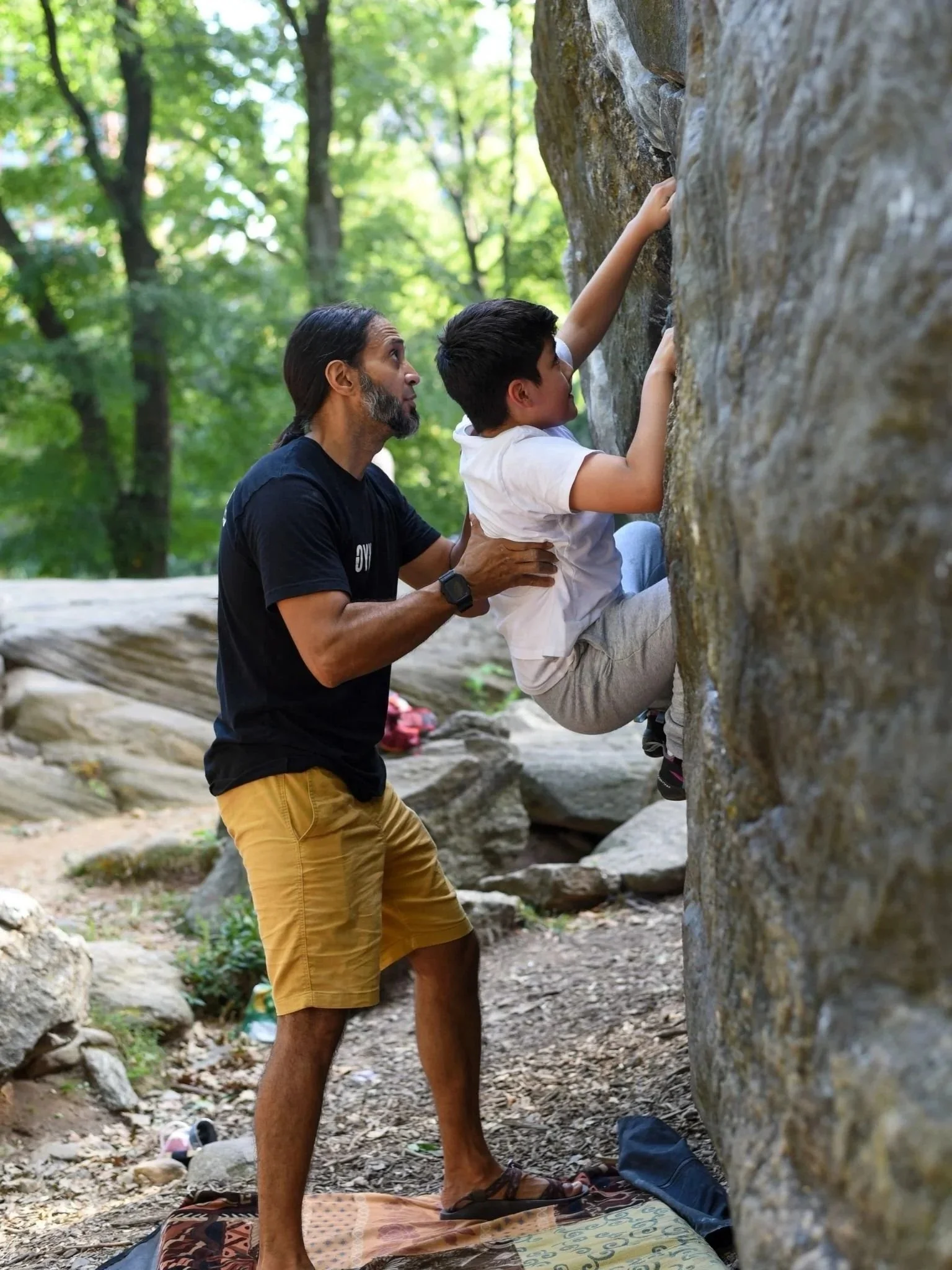 A mentor spots a young boy as he climbs a large boulder in a wooded park.