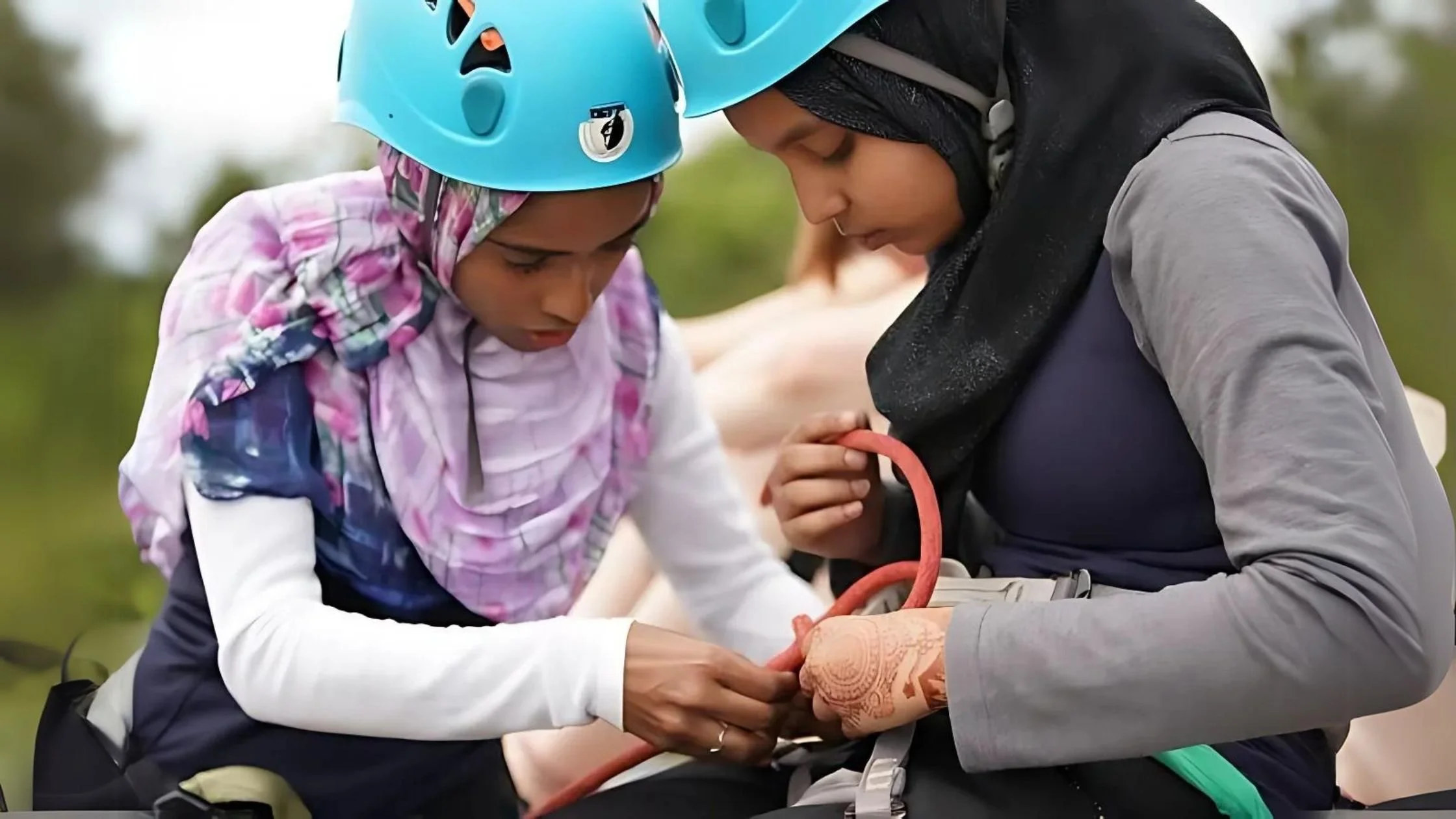 Two women, both wearing helmets and religious headscarves, one woman has detailed henna on her hand, collaborate to secure a climbing rope, focusing on safety techniques.