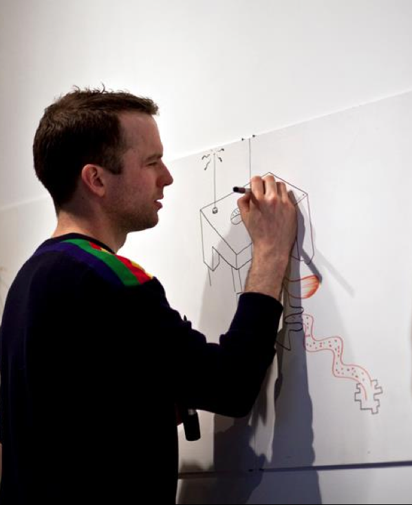 A man drawing a cartoon robot on a white wall with a black marker, casting a shadow on the wall.