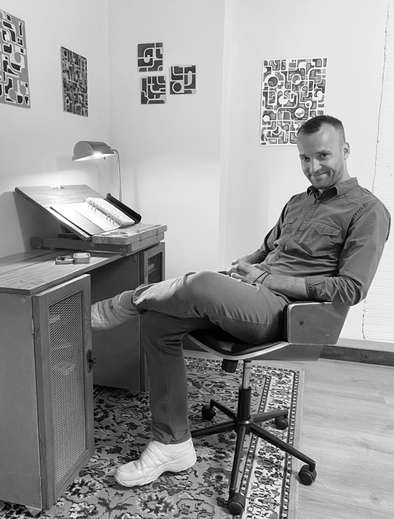 Man sitting at a desk in a room with modern art on the walls and a patterned rug, smiling at the camera.
