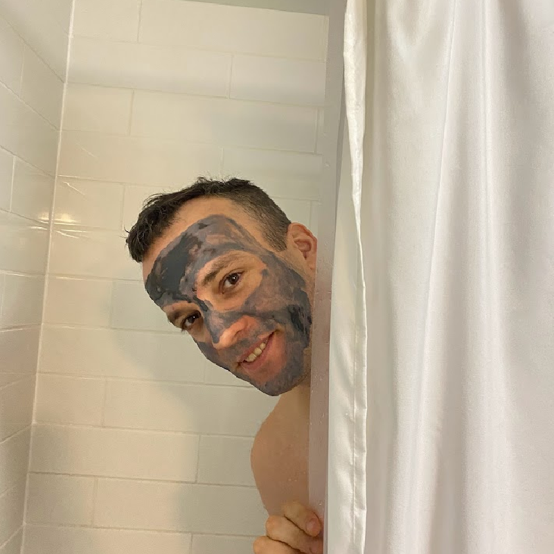 A man smiling in the shower with a charcoal face mask, peeking out from behind a shower curtain.