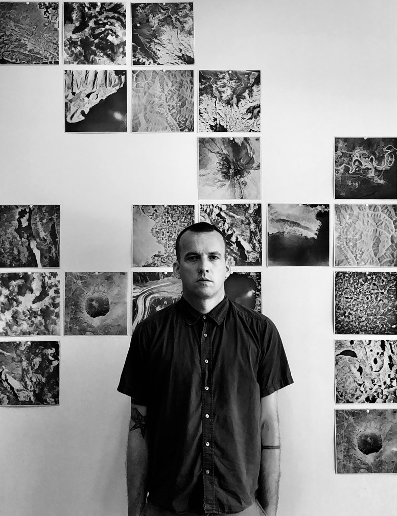 A man with short hair and tattoos on his arms stands in front of a wall with various black and white satellite images or maps displayed in a grid pattern.