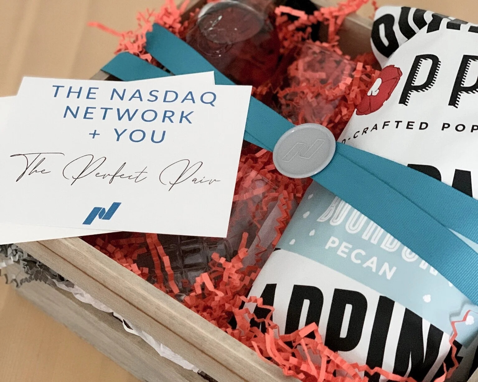 A gift box containing a card with the NASDAQ logo and the message 'The NASDAQ Network + You', a bag of artisanal popcorn, and a bottle of beverage. The gift is decorated with shredded paper filling.
