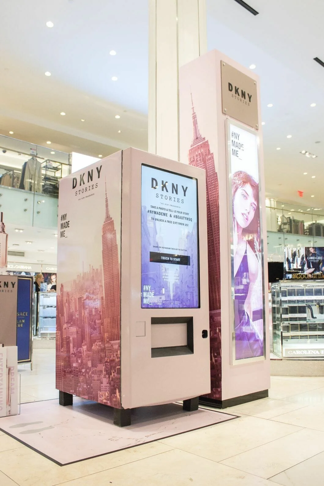 A photo booth in a shopping mall with pink and purple Empire State Building graphics and digital screens promoting DKNY Stories and a campaign called #NYMadeMe. The booth has a digital display instructing users to take a photo and share it on Instagram to unlock a free gift.