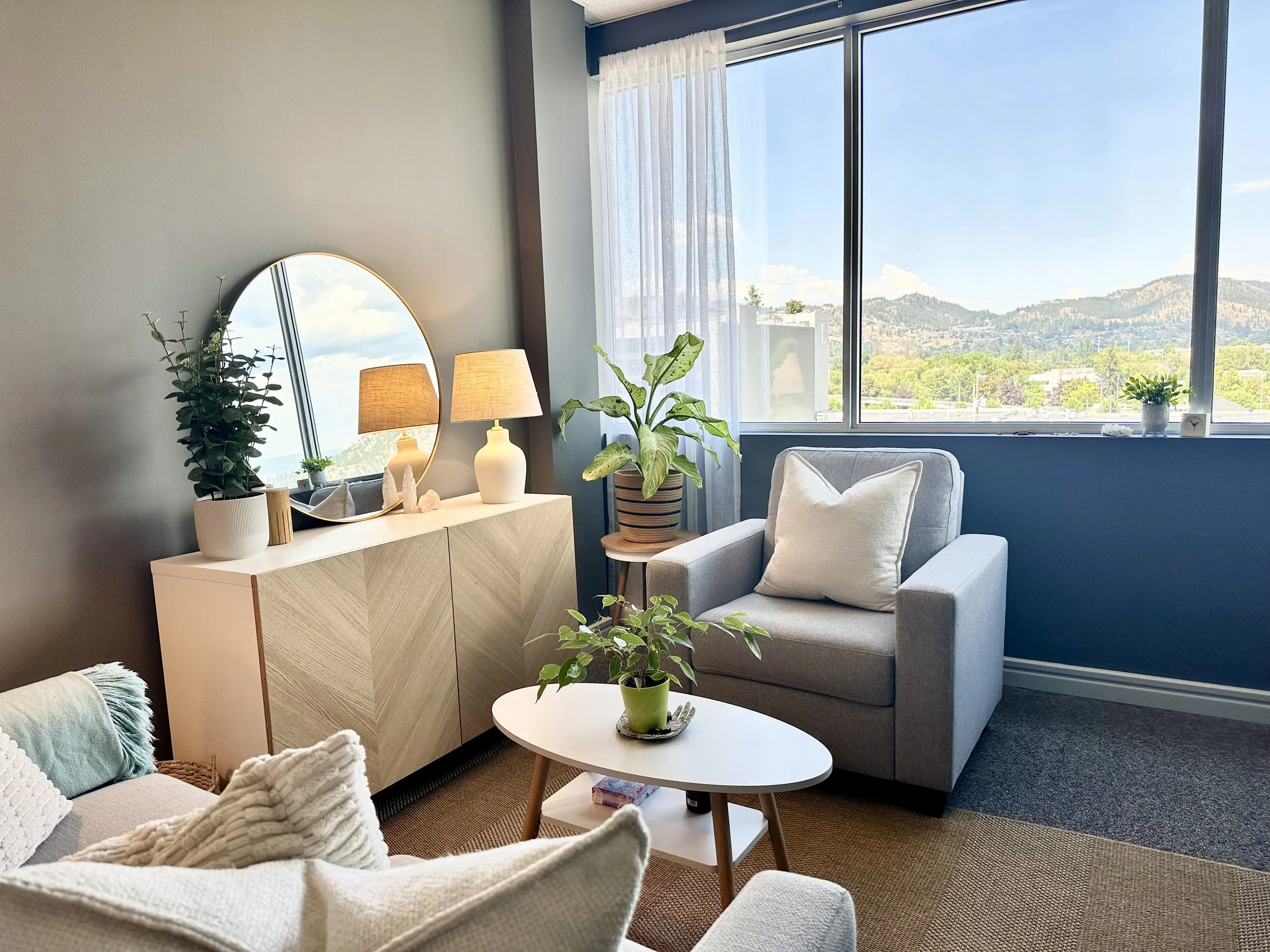 Cozy counselling office with large window showing a mountain view, gray armchair with pillow, white sideboard with plants and lamps, round mirror, and a coffee table with a potted plant.
