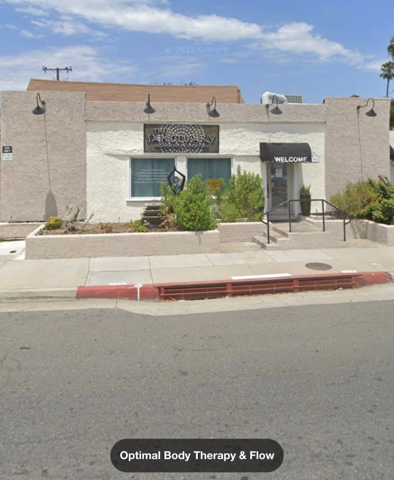 Exterior of a building with a sign that says 'The Sanctuary' and a black awning with 'Welcome' written on it. There are windows, plants, and stairs leading to the entrance. The image is captioned 'Optimal Body Therapy & Flow'.