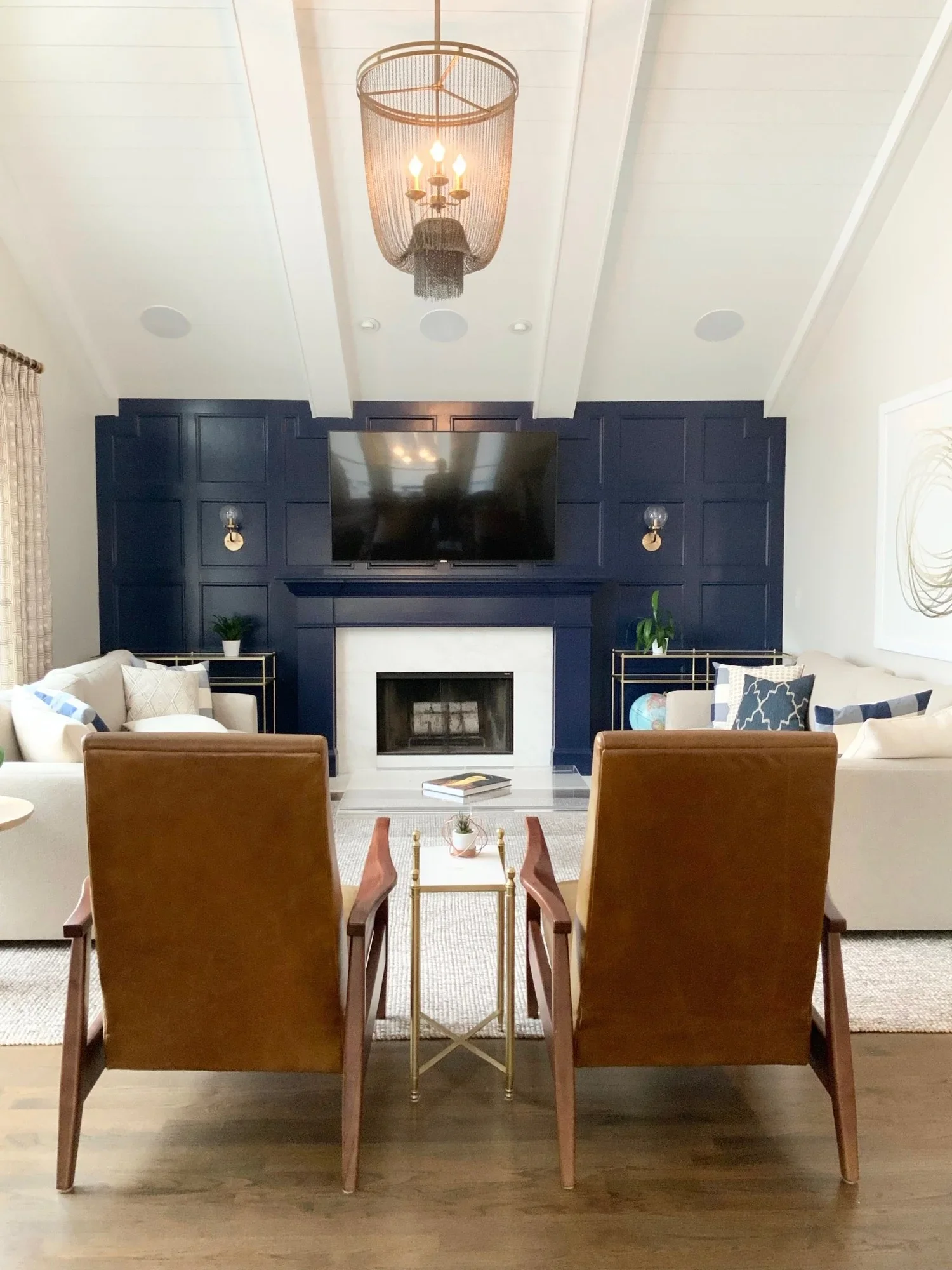 Living room with a fireplace surrounded by navy blue paneling, a large flat-screen TV, and a chandelier. There are two beige couches flanking the fireplace, and two coffee tables with decor. The ceiling is white with built-in lighting and a central c