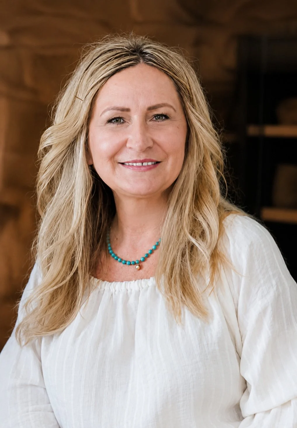 A woman with long blonde hair, wearing a white blouse and a turquoise beaded necklace, smiling at the camera. She is indoors with a wooden background.