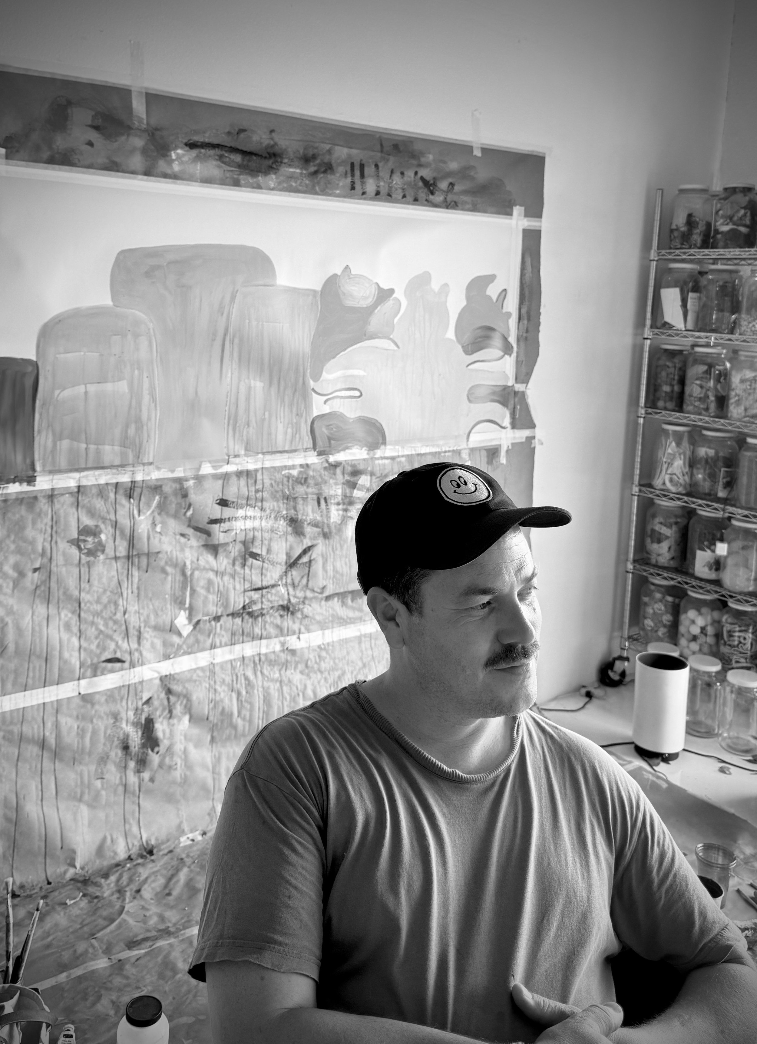 A man with a mustache wearing a cap with a smiley face logo, sitting at a table in a room with shelves of jars and containers, and a large artwork of mushrooms on the wall behind him.