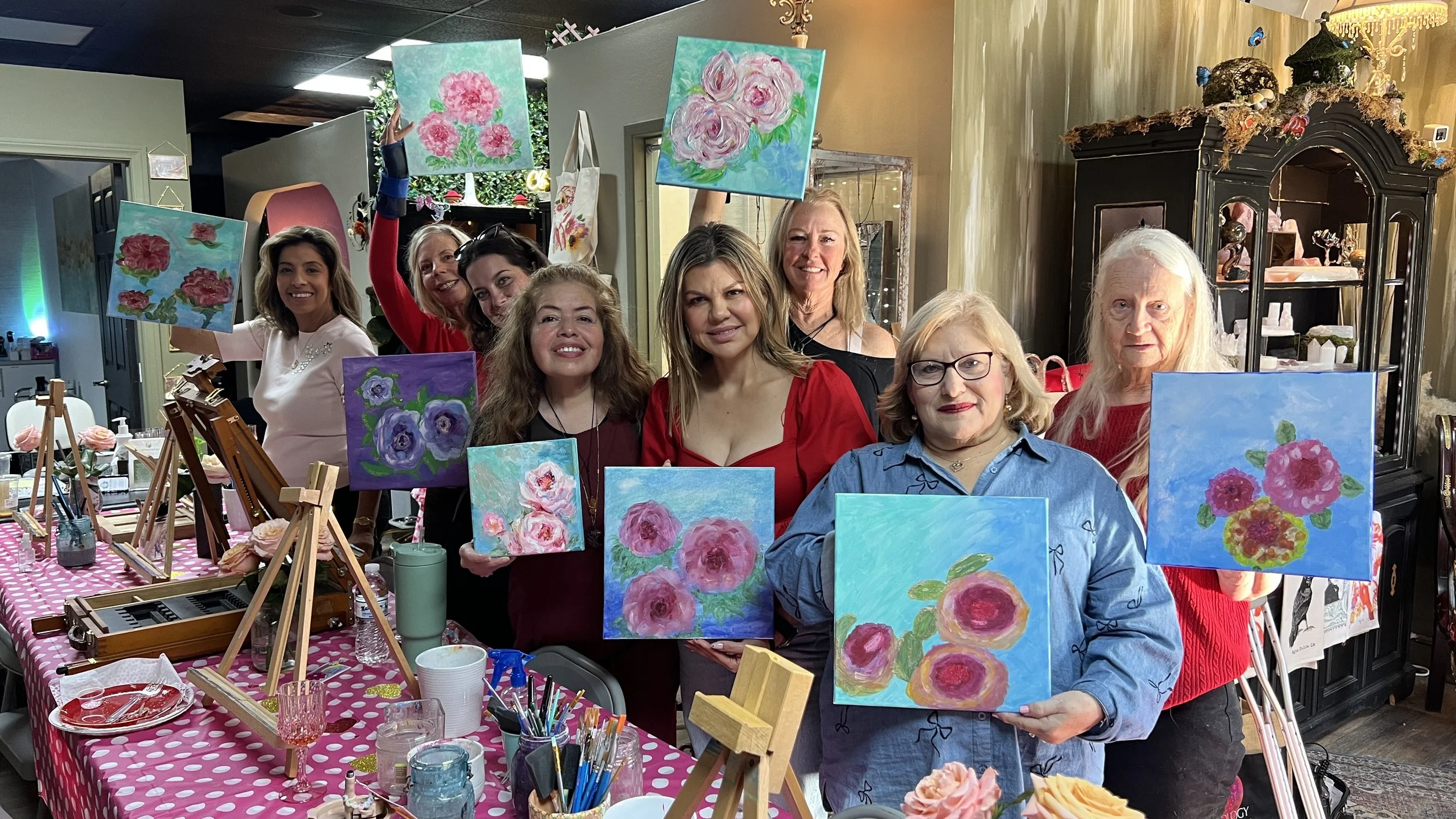 A group of women holding up paintings of pink and purple flowers, displaying their artwork at a floral painting class or gathering.