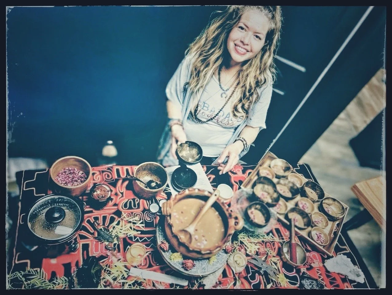 Young woman smiling at a table with various bowls and cups, preparing a traditional drink, possibly chocolate with toppings, and surrounded by flowers or decorations.