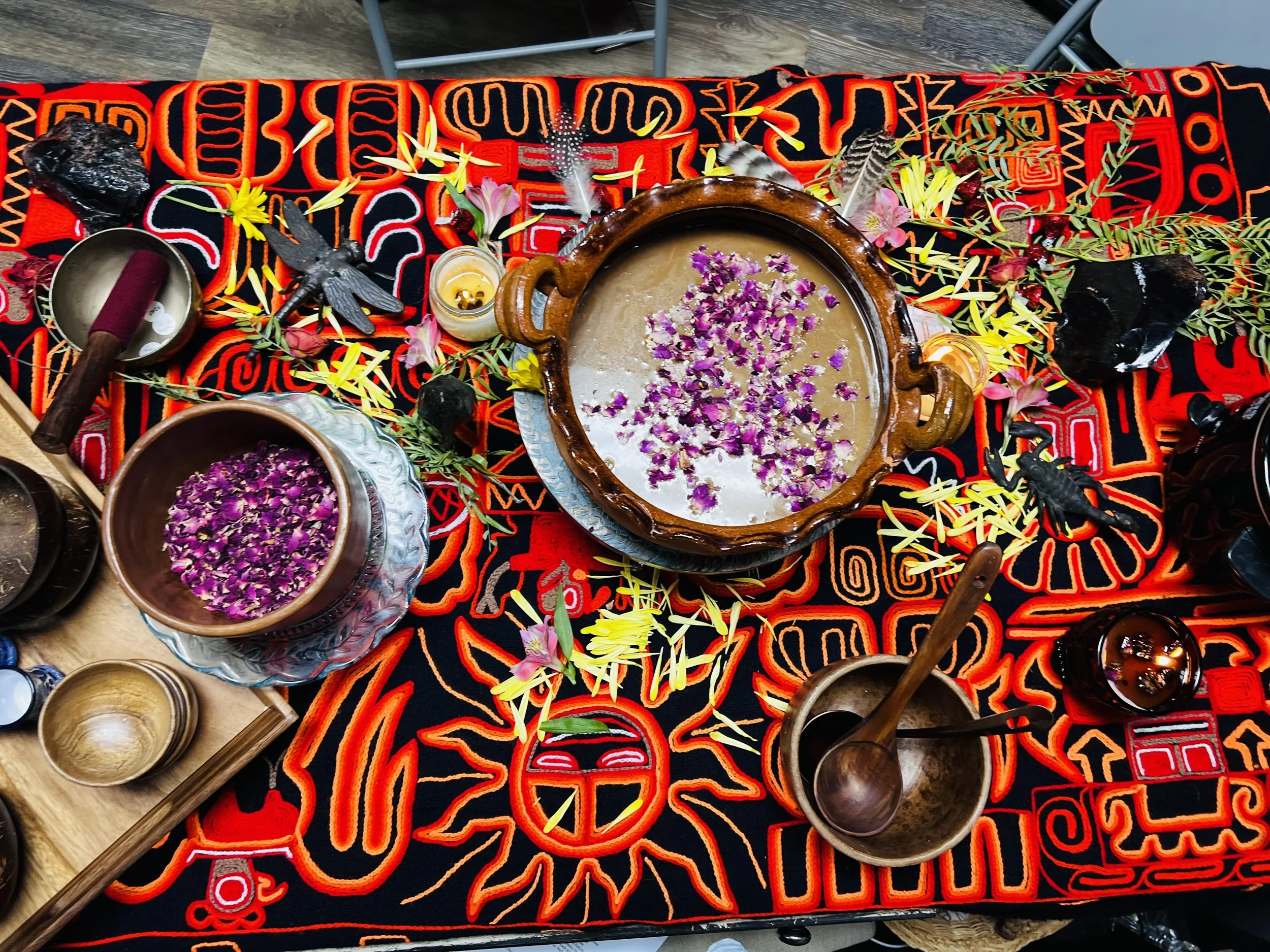 A decorated table with flowers, feathers, candles, bowls, and a large clay vessel with a floral pattern, set on colorful embroidered fabric.